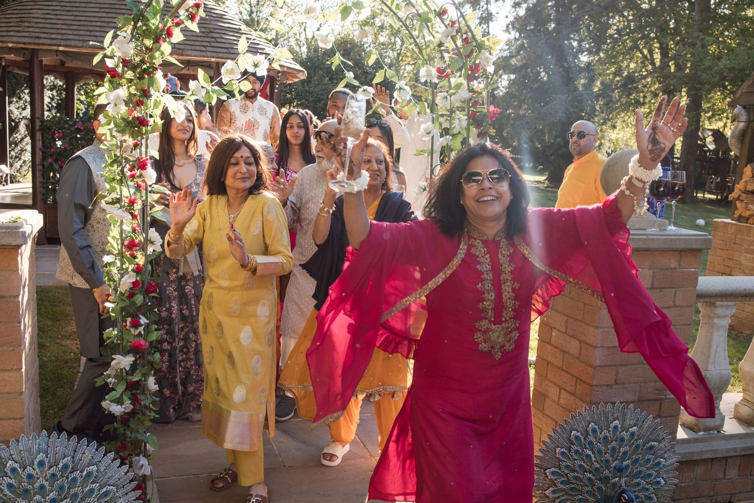 Wedding guests in vibrant Indian attire walk through a floral garden archway during a festive procession.