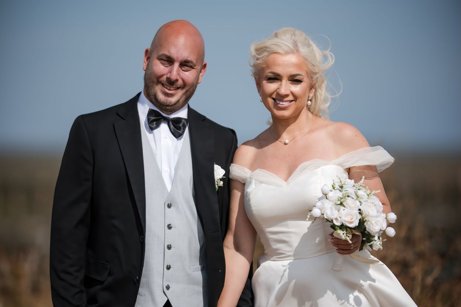 Smiling bride and bald groom pose together outdoors, bride holding white floral bouquet.