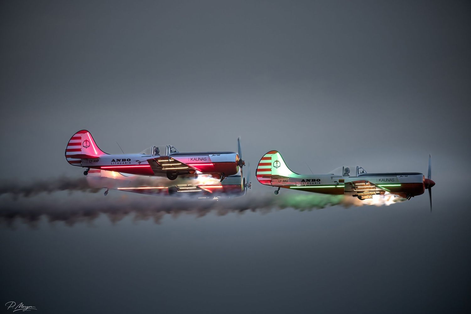 Two colorful propeller aircraft perform a low-altitude airshow formation flight trailing colorful smoke.