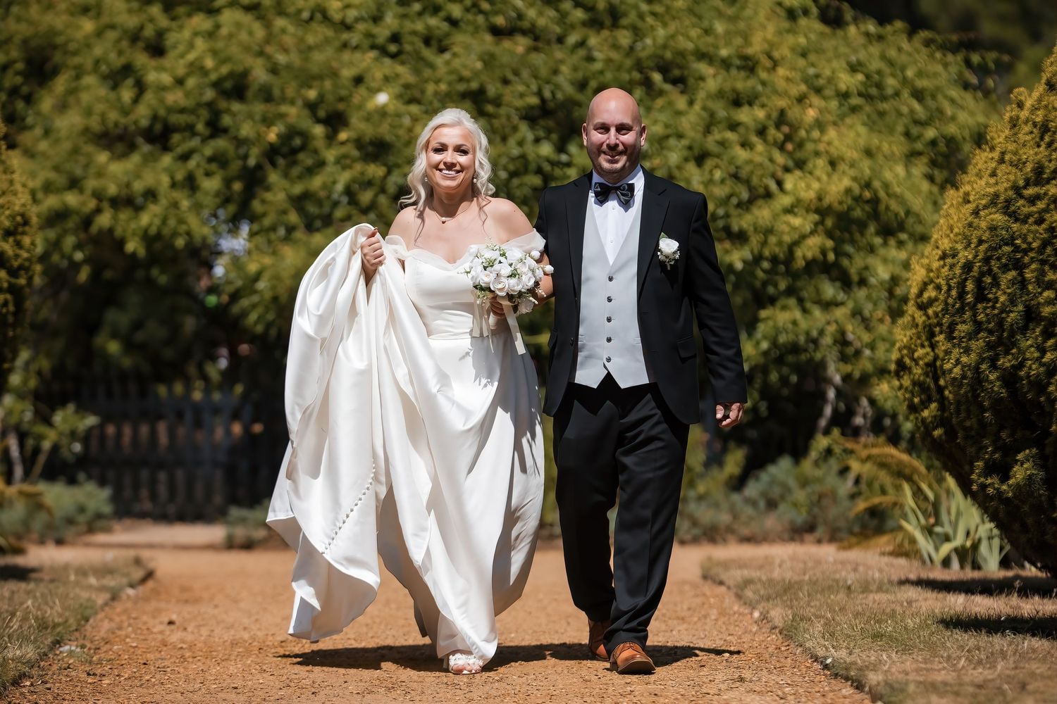 Happy newlyweds walking hand in hand along sunny garden path, bride carrying white bouquet in elegant gown.