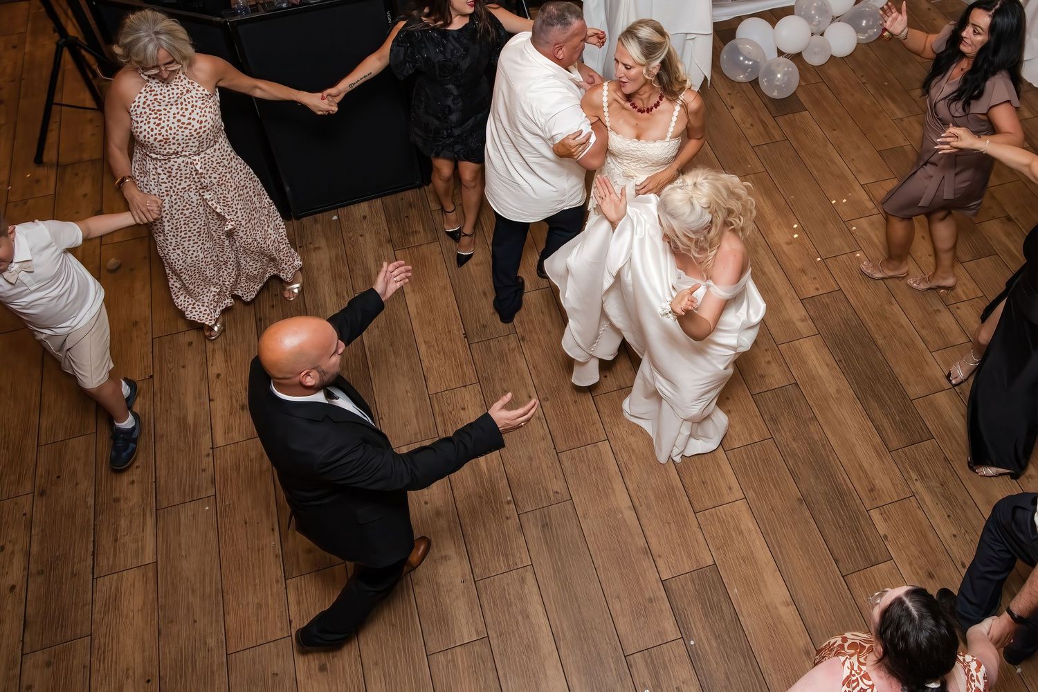 Aerial view of bride and groom dancing joyfully with wedding guests on wooden reception dance floor.