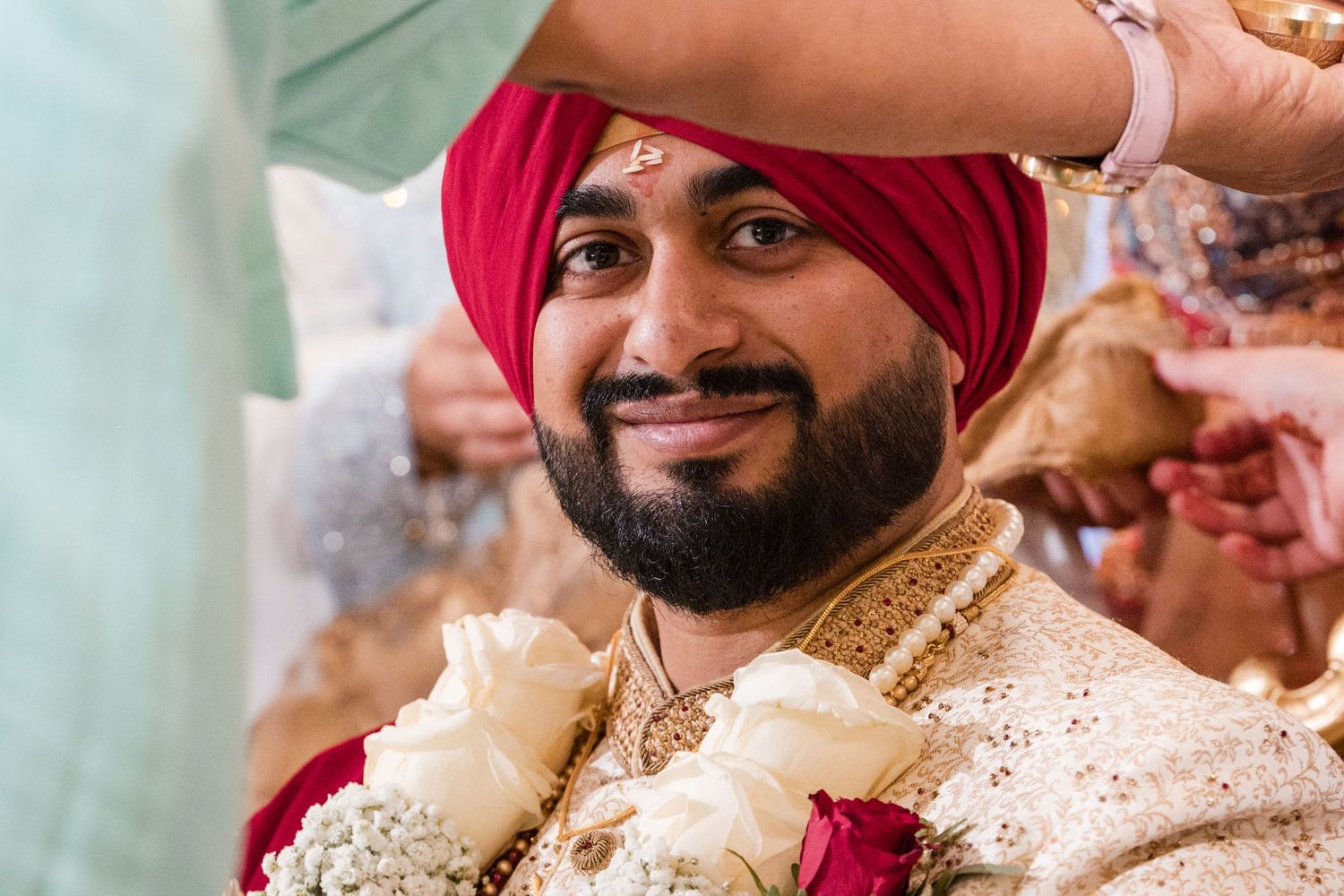 Sikh groom smiling wearing red turban and floral garland at traditional wedding ceremony.