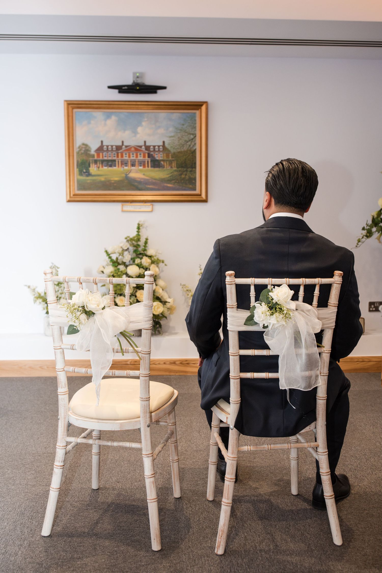 Groom in dark suit sits alone at decorated chiavari chairs awaiting bride at intimate wedding ceremony.