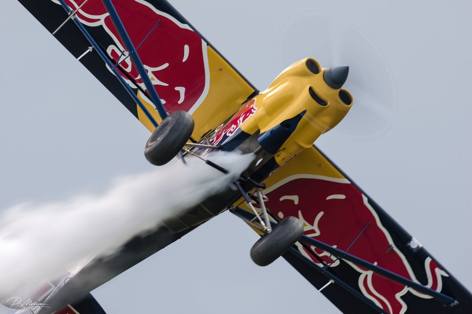 A Red Bull aerobatic plane with bold logo performs a dramatic maneuver leaving a smoke trail.