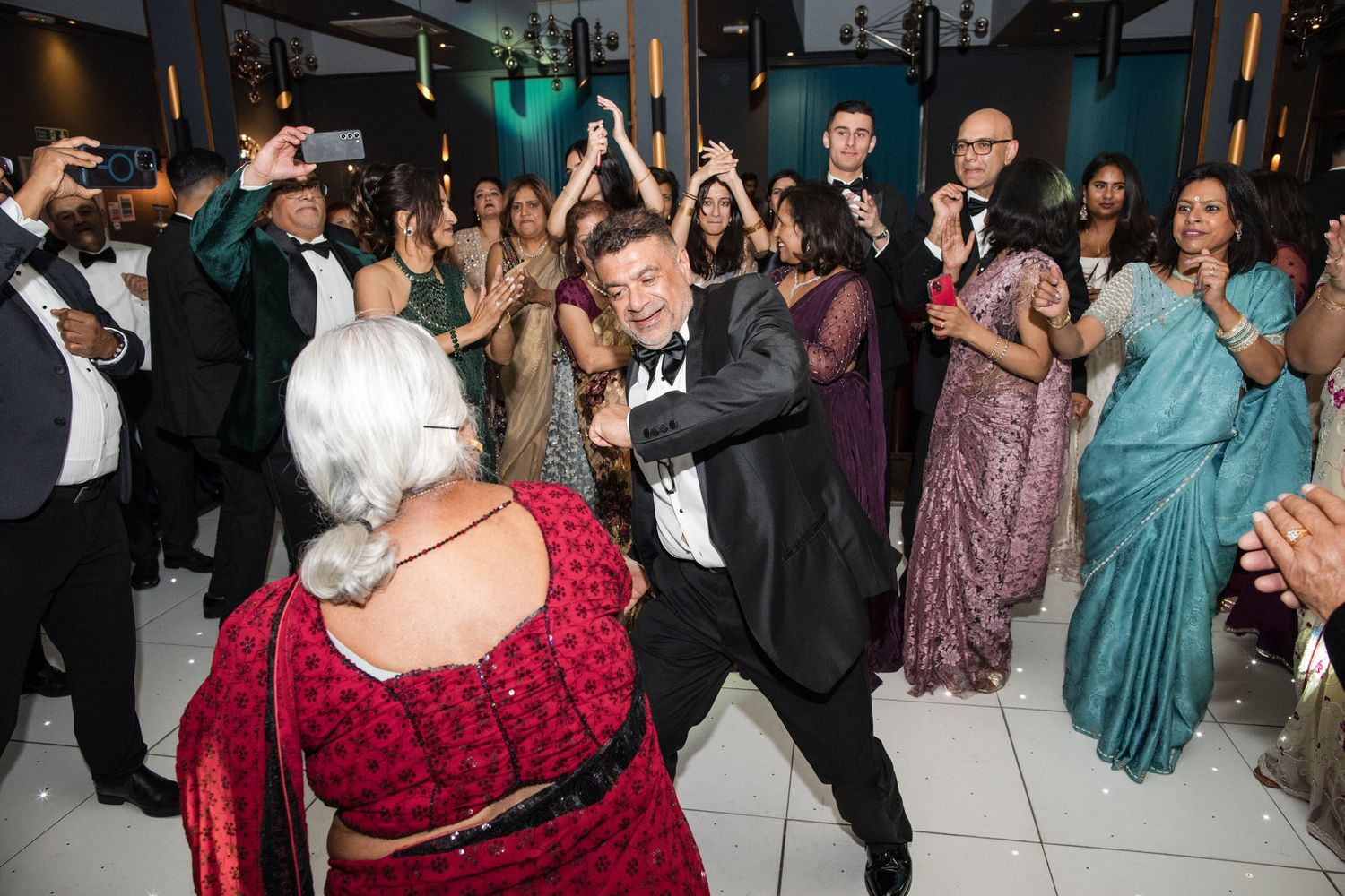 Groom in tuxedo dancing energetically with elderly guest in red dress at Indian wedding reception.