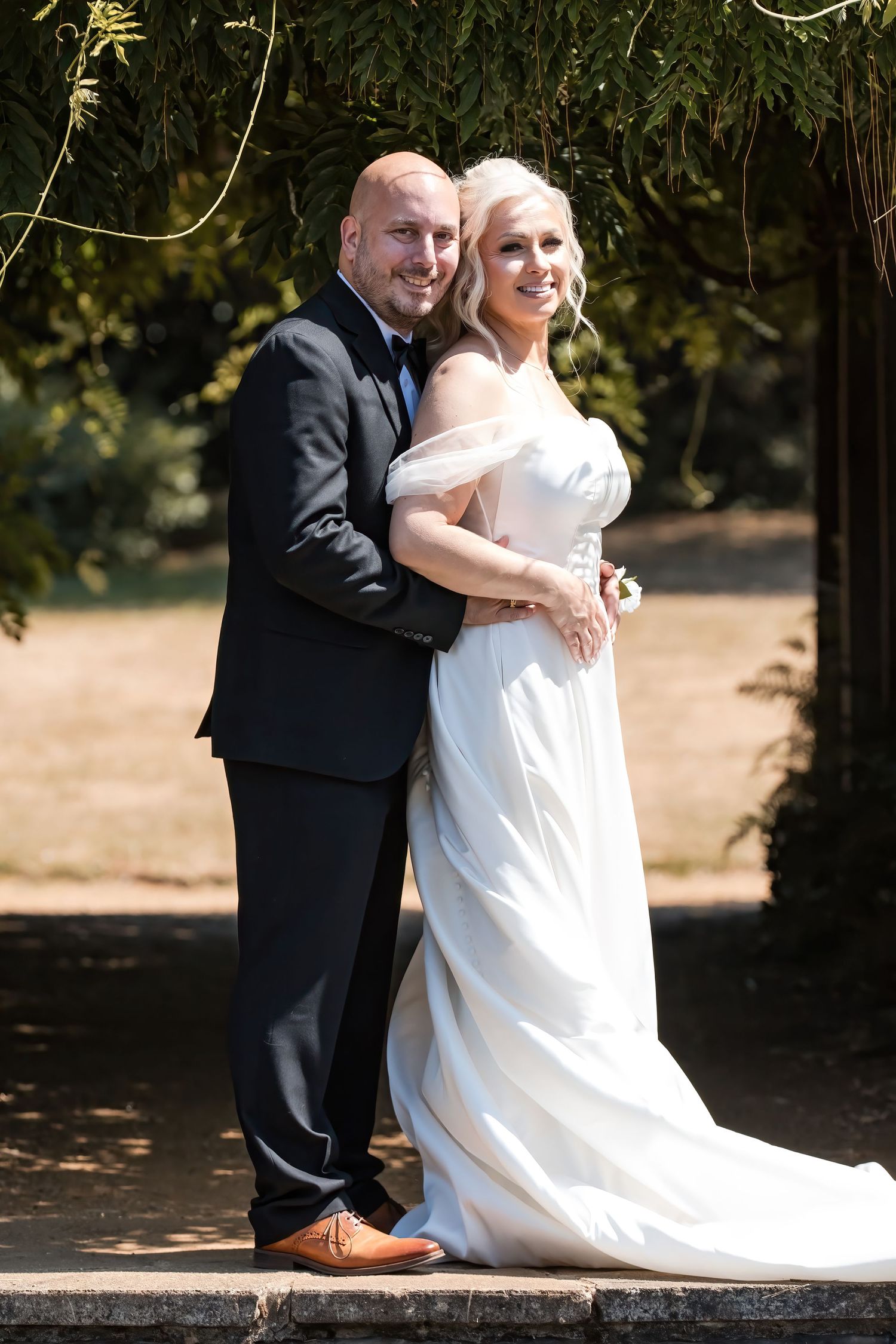 Elegant wedding couple posing together under green foliage, groom in black suit embracing bride in white gown.
