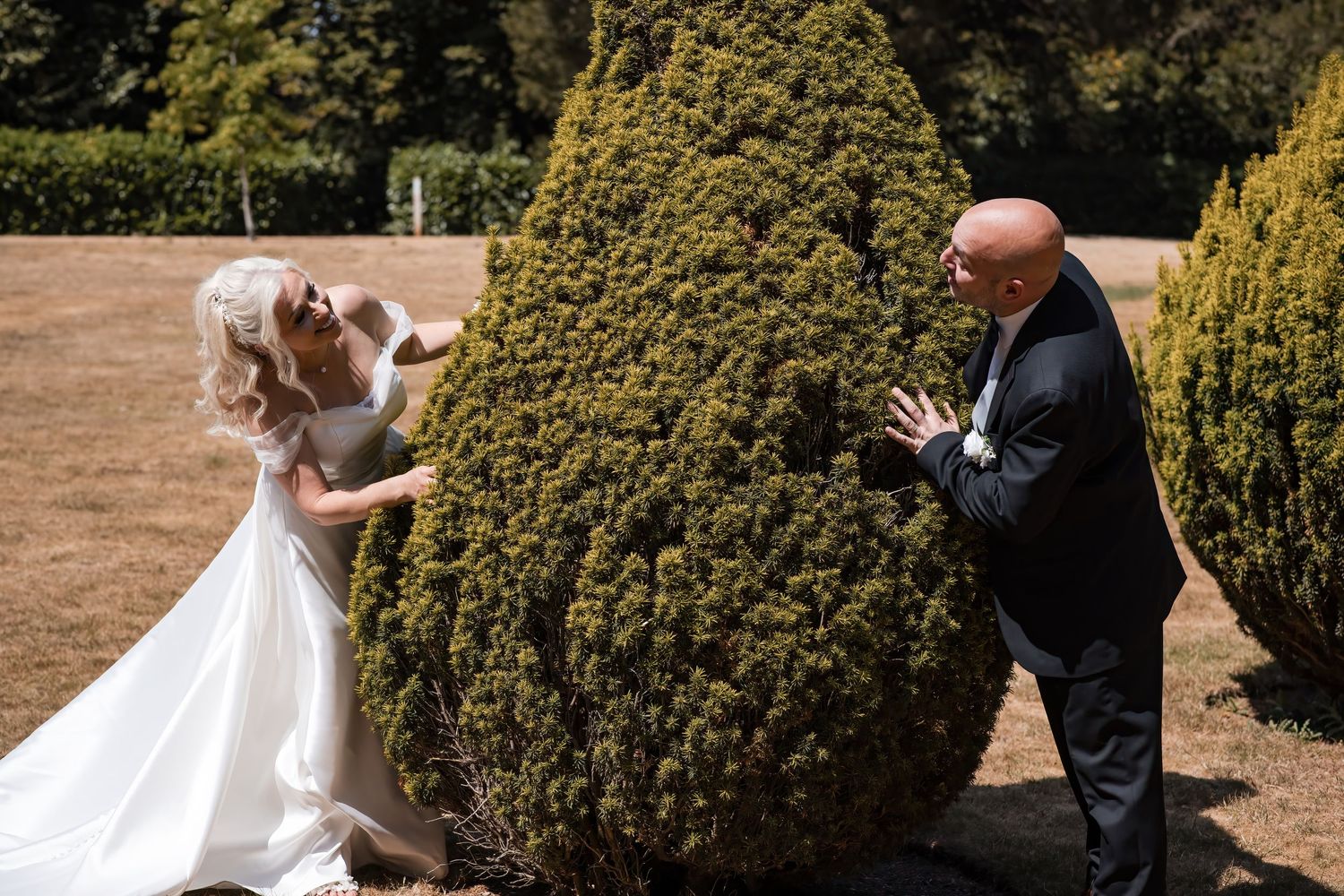 Bride and groom share a playful moment hiding behind a large topiary bush in a garden.