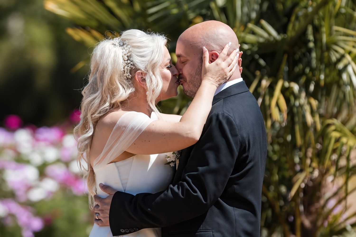 Bride and bald groom share first kiss embracing passionately in front of vibrant pink garden flowers.