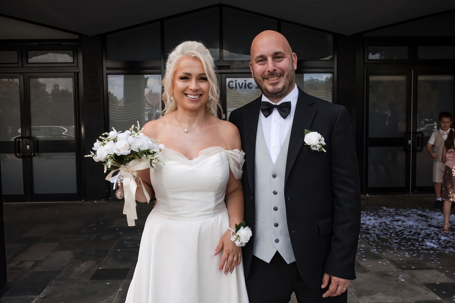 Smiling bride and groom pose outside civic wedding venue, bride holding white bouquet in elegant off-shoulder gown.
