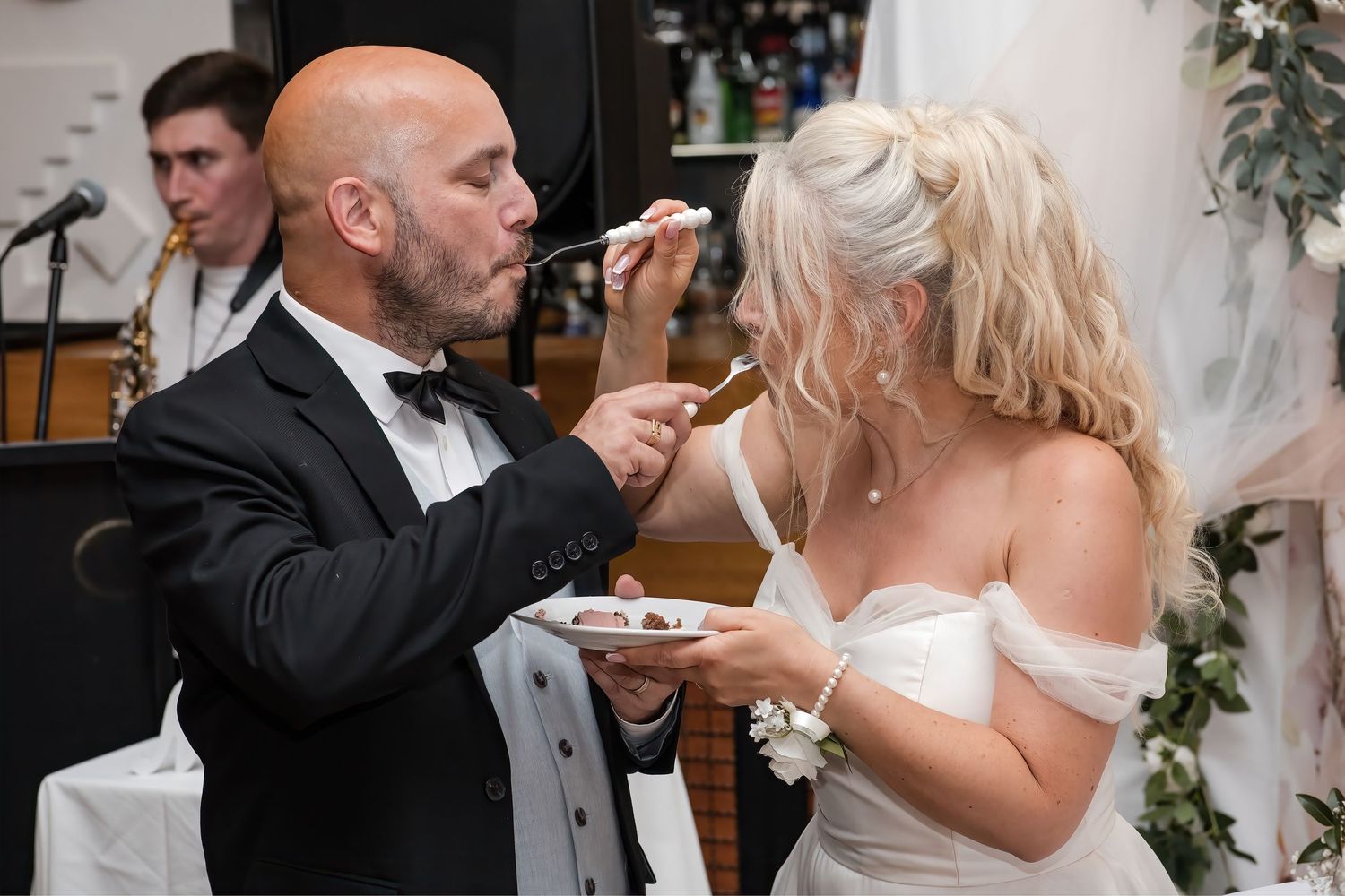 Bride and groom playfully feed each other wedding cake at elegant white-decorated reception venue.