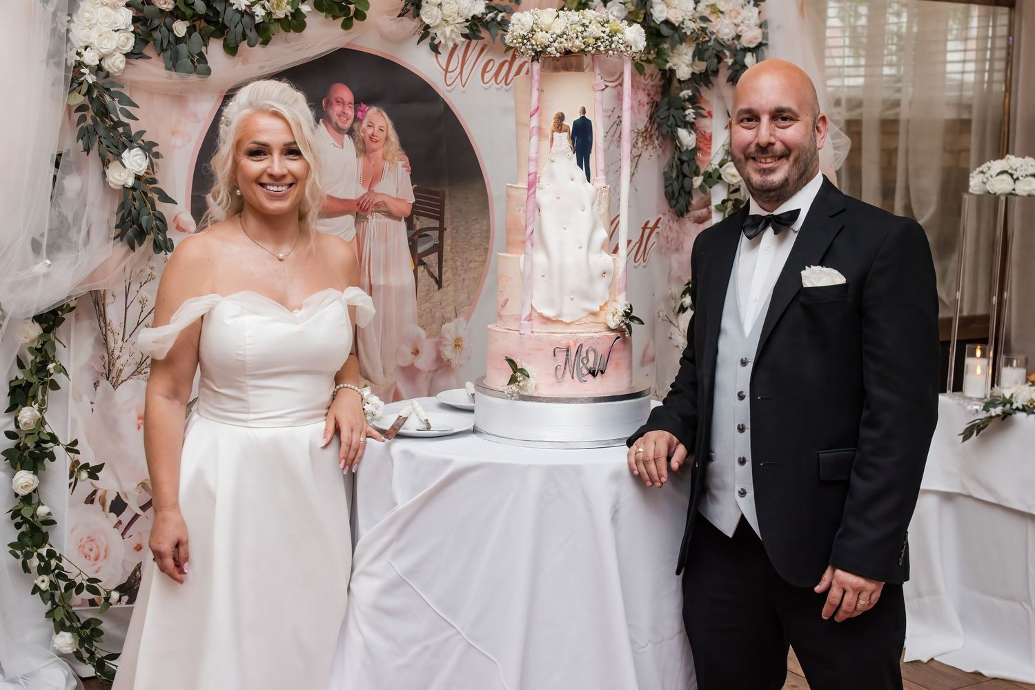 Newlyweds pose beside their elegant multi-tiered wedding cake decorated with floral accents.