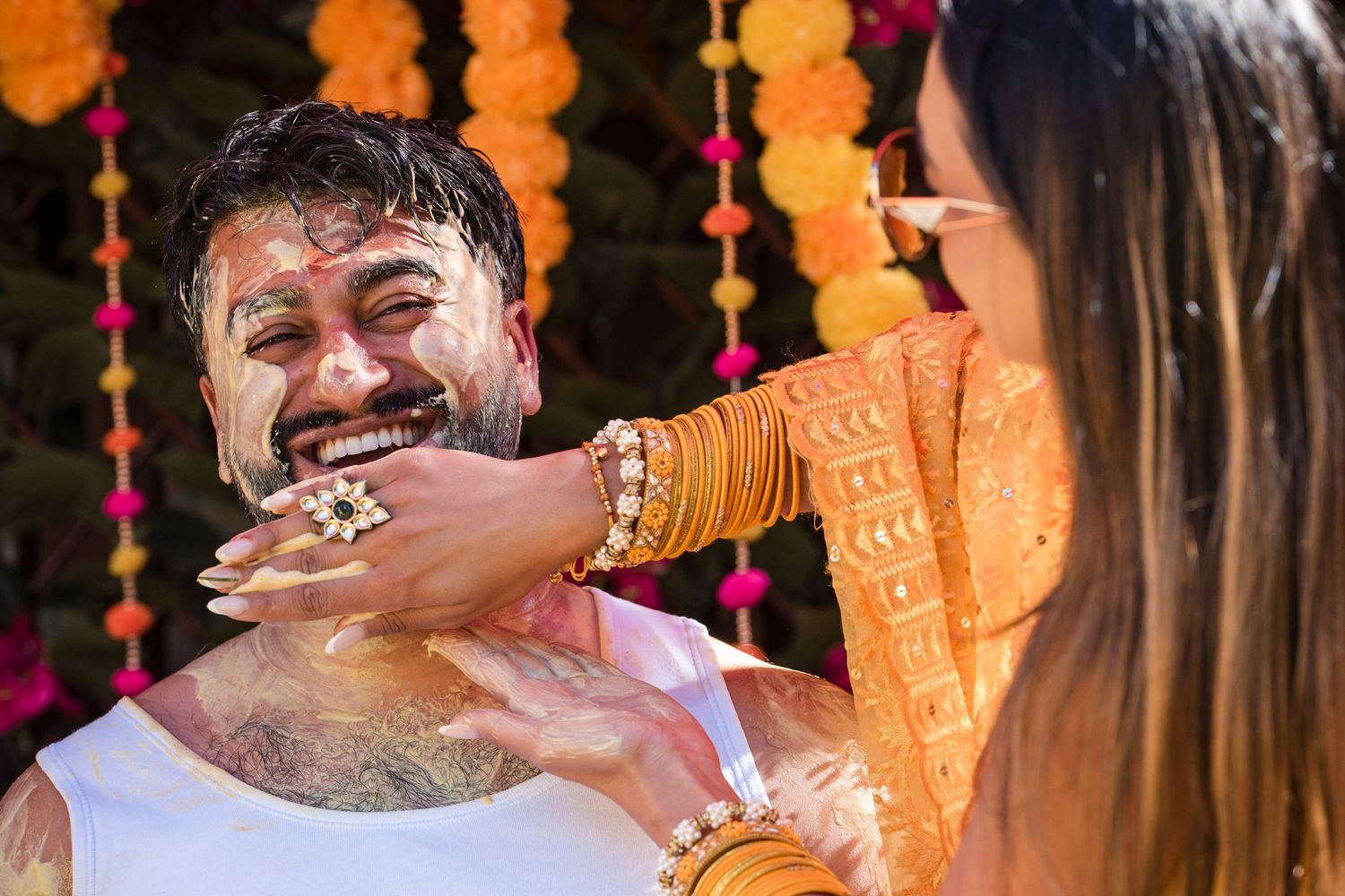 Joyful groom laughing as bride playfully applies haldi paste during colorful Indian pre-wedding ceremony.