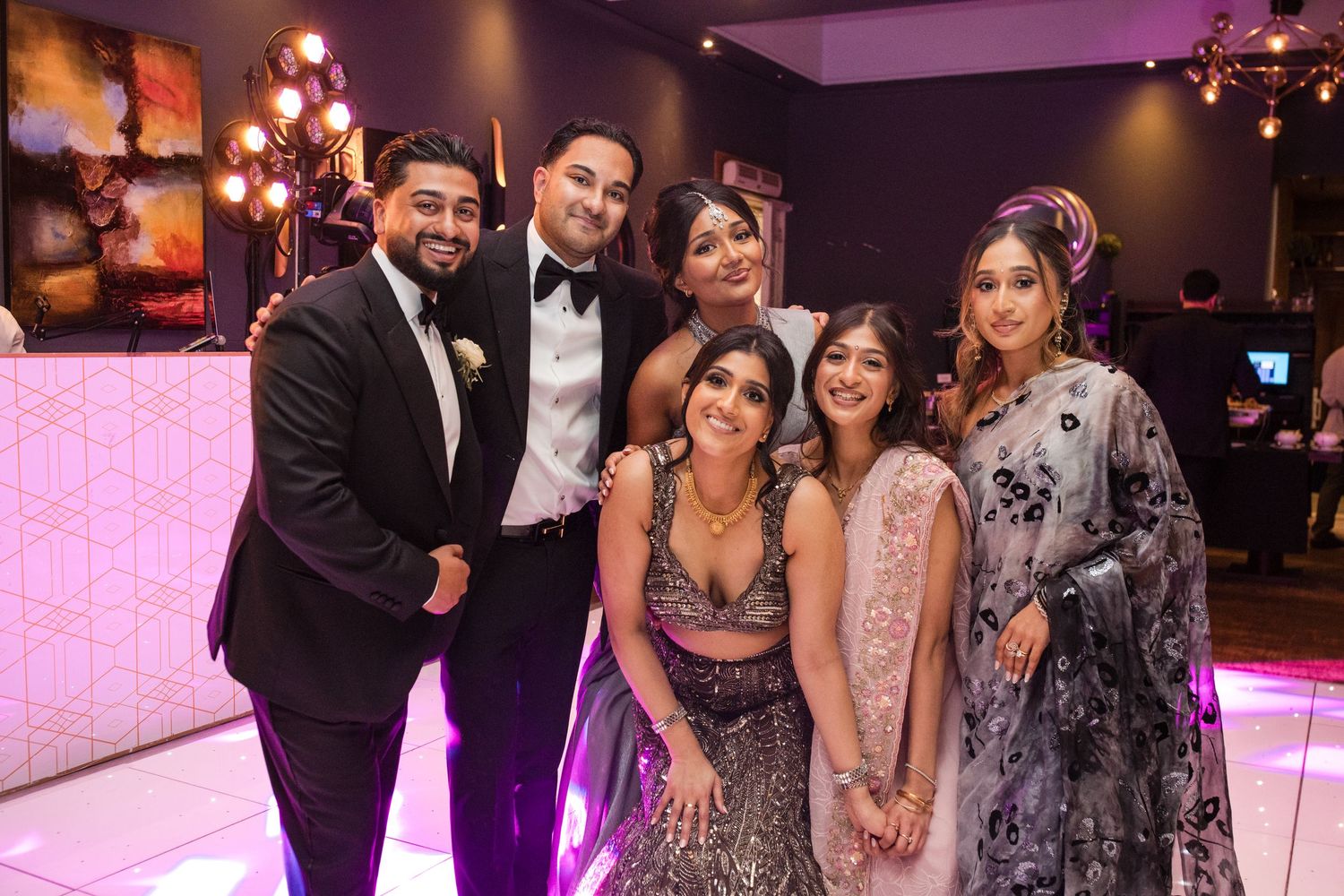Group of six friends posing together and smiling at an Indian wedding reception under pink lights.