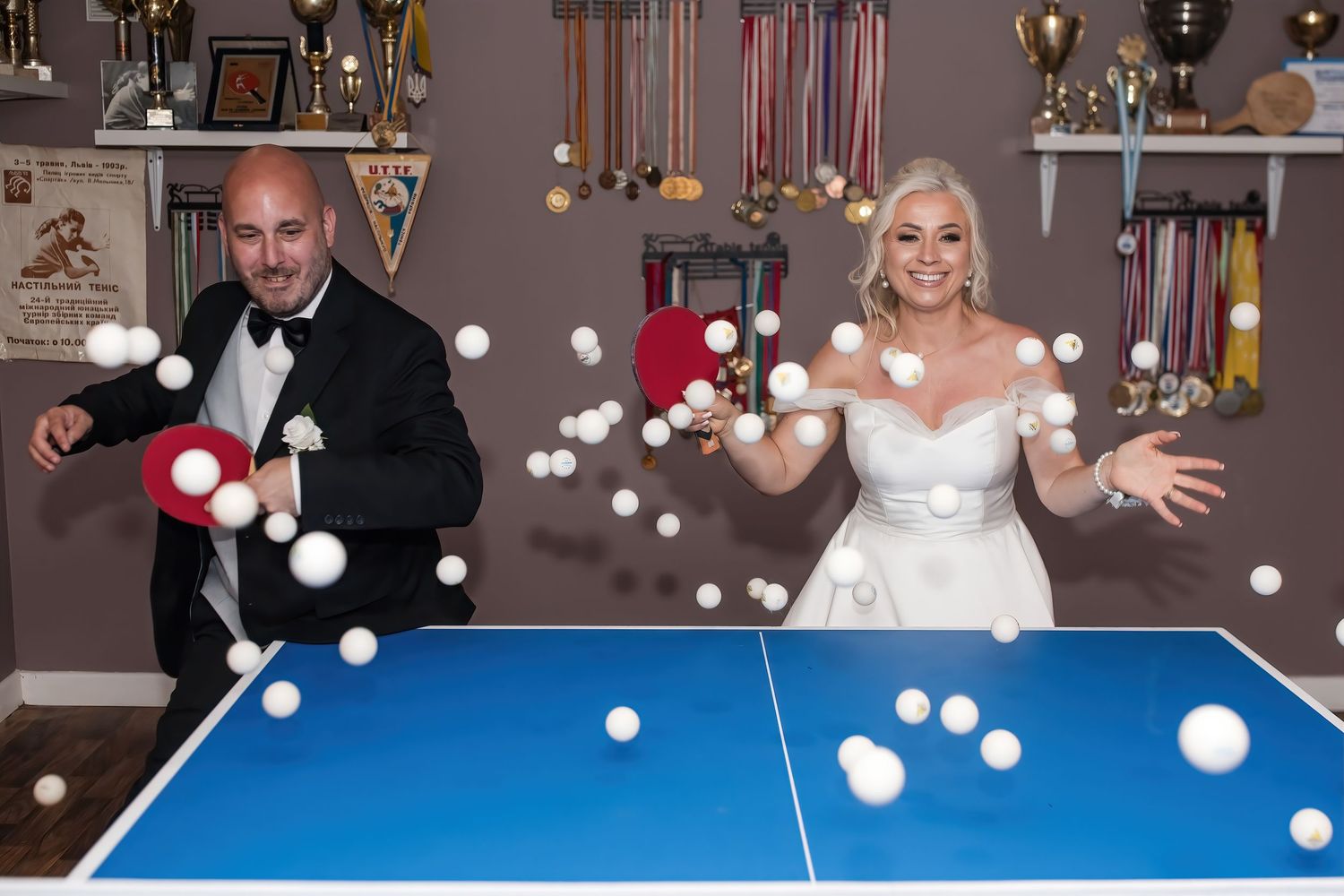 Bride and groom laugh while playing ping pong with balls flying across a blue table.