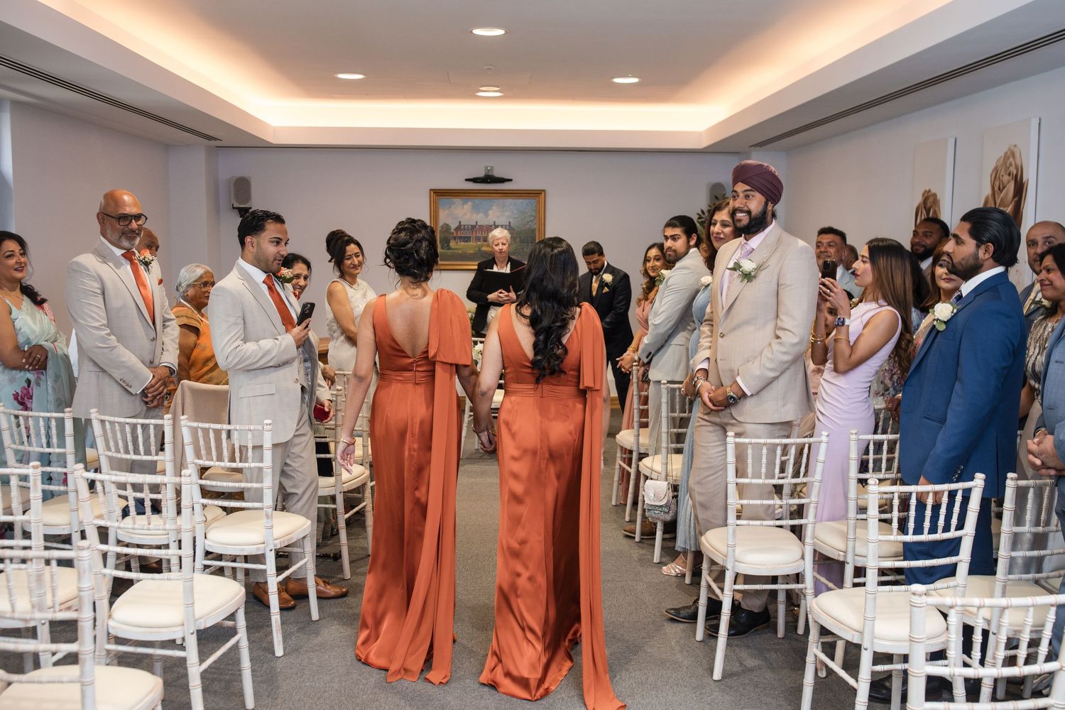 Bride in white gown walks down aisle with bridesmaids in rust dresses at modern wedding ceremony.