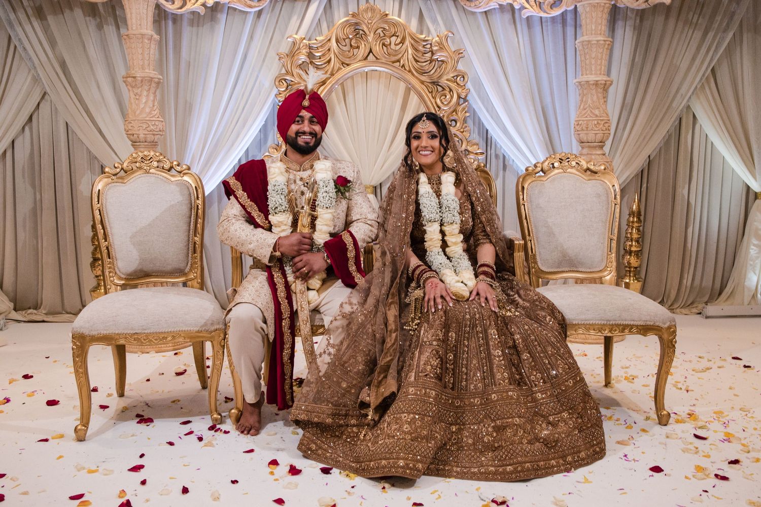 Sikh bride and groom in traditional attire seated at ornate wedding mandap with floral decorations.