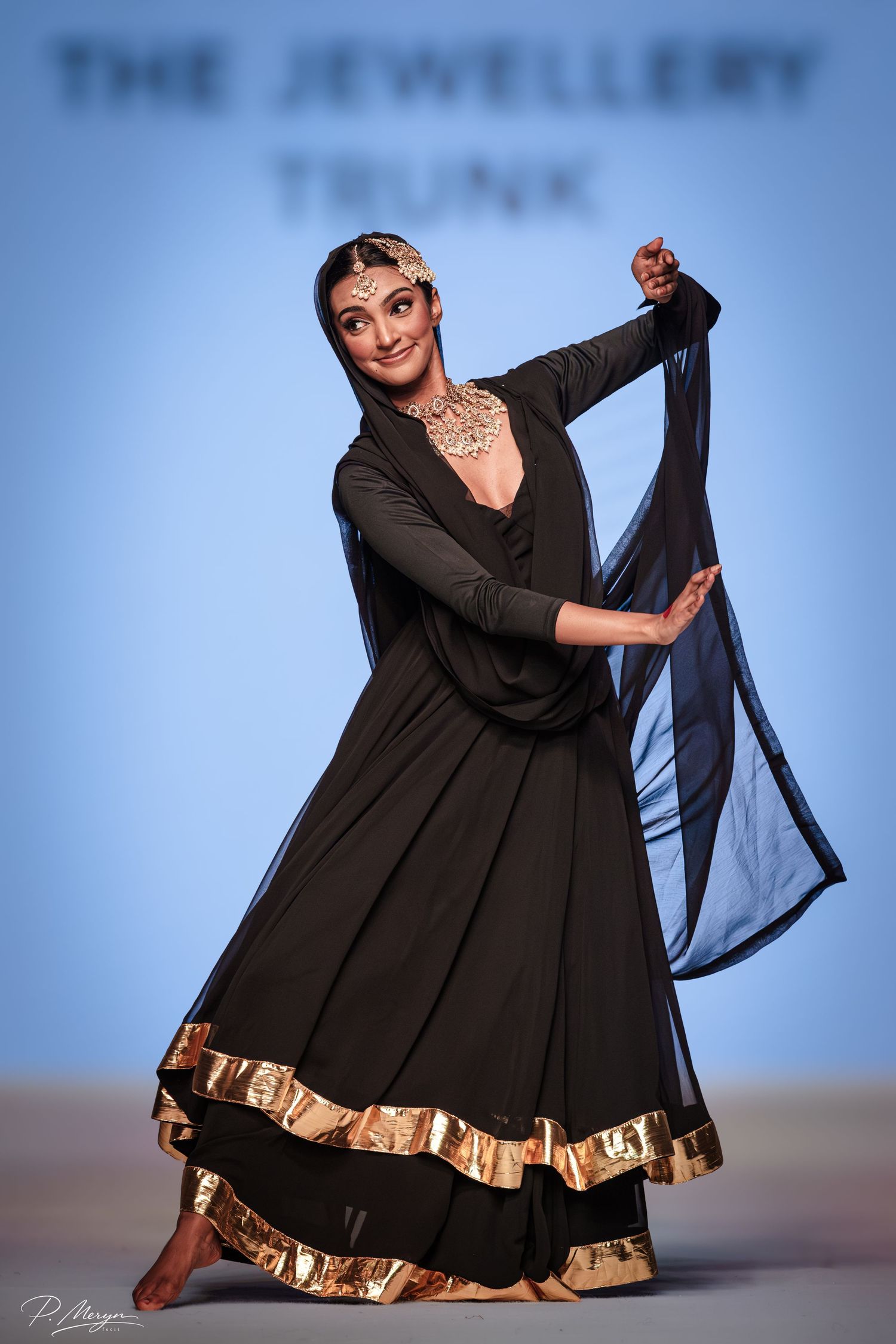 Dancer in black anarkali with gold trim performs graceful pose on runway at The Jewellery Trunk show.