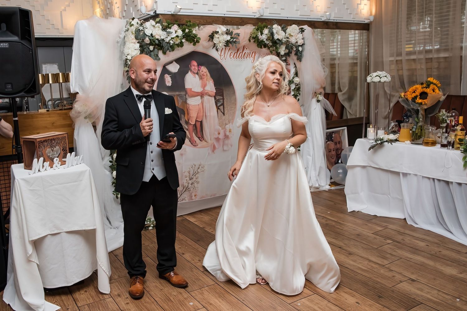 Bride and groom address wedding guests during reception speech in beautifully decorated venue with floral backdrop.