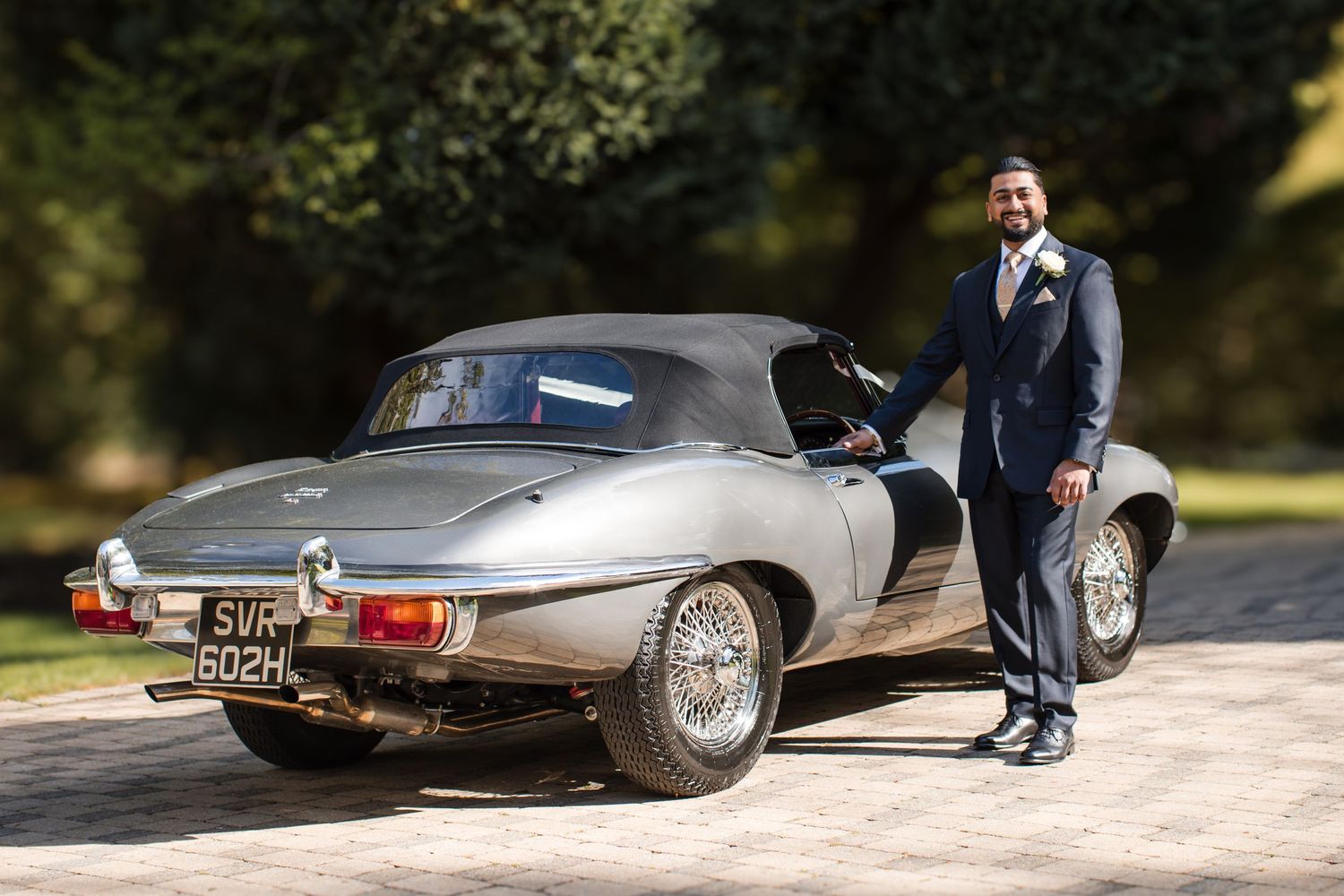 Dapper groom in navy suit posing beside a classic vintage Jaguar E-Type sports car outdoors.