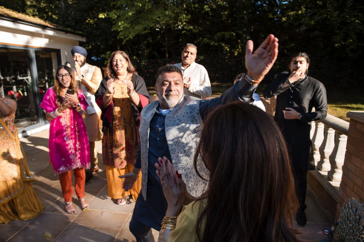 Family and friends in traditional South Asian clothing celebrate joyfully on a sunlit outdoor patio.