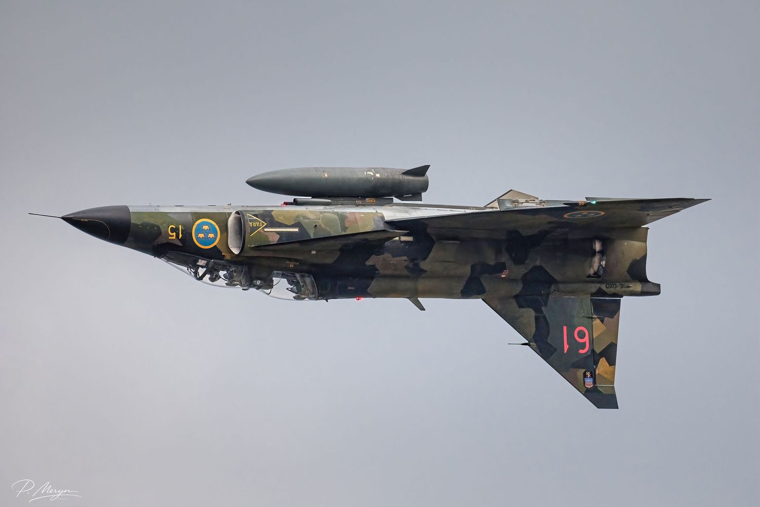 A camouflaged Swedish Saab Viggen fighter jet numbered 19 climbs steeply during an airshow.