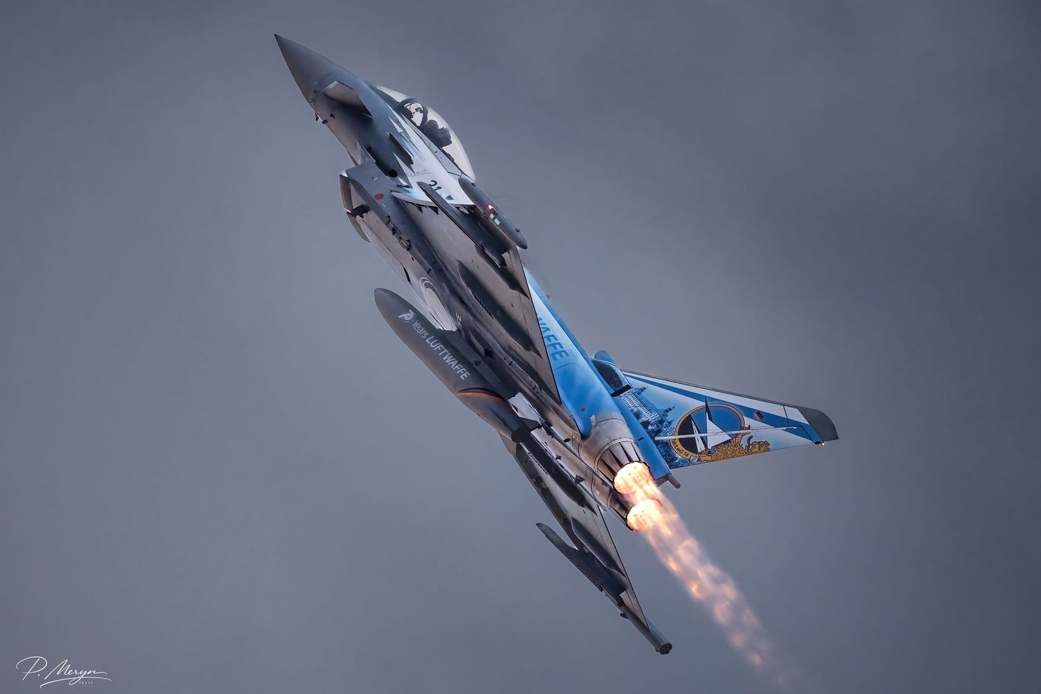 Modern F-15 fighter jet with blue livery climbing steeply with afterburner flames during airshow performance.