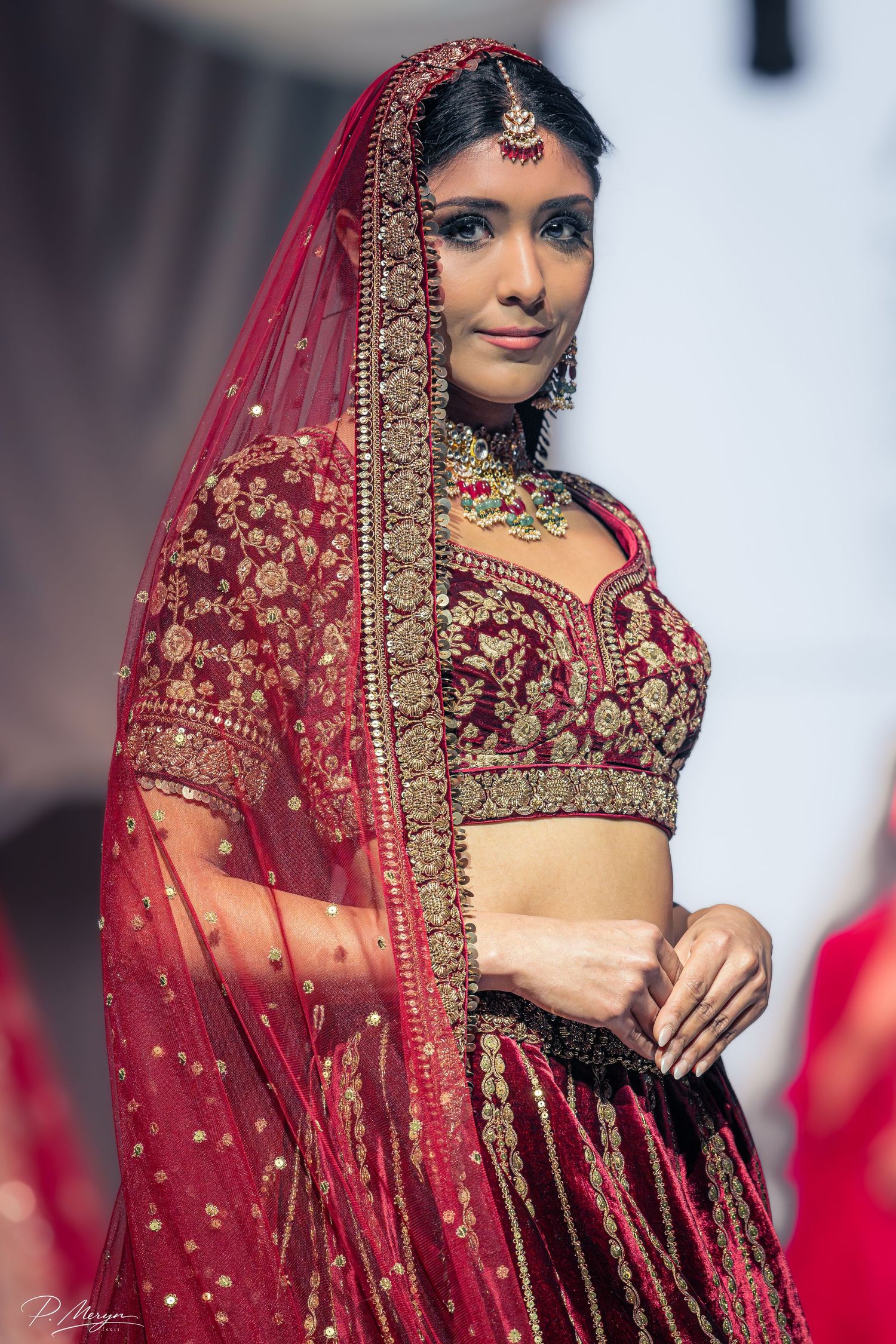 Model wearing red bridal lehenga with gold embroidery and matching dupatta at South Asian fashion show.