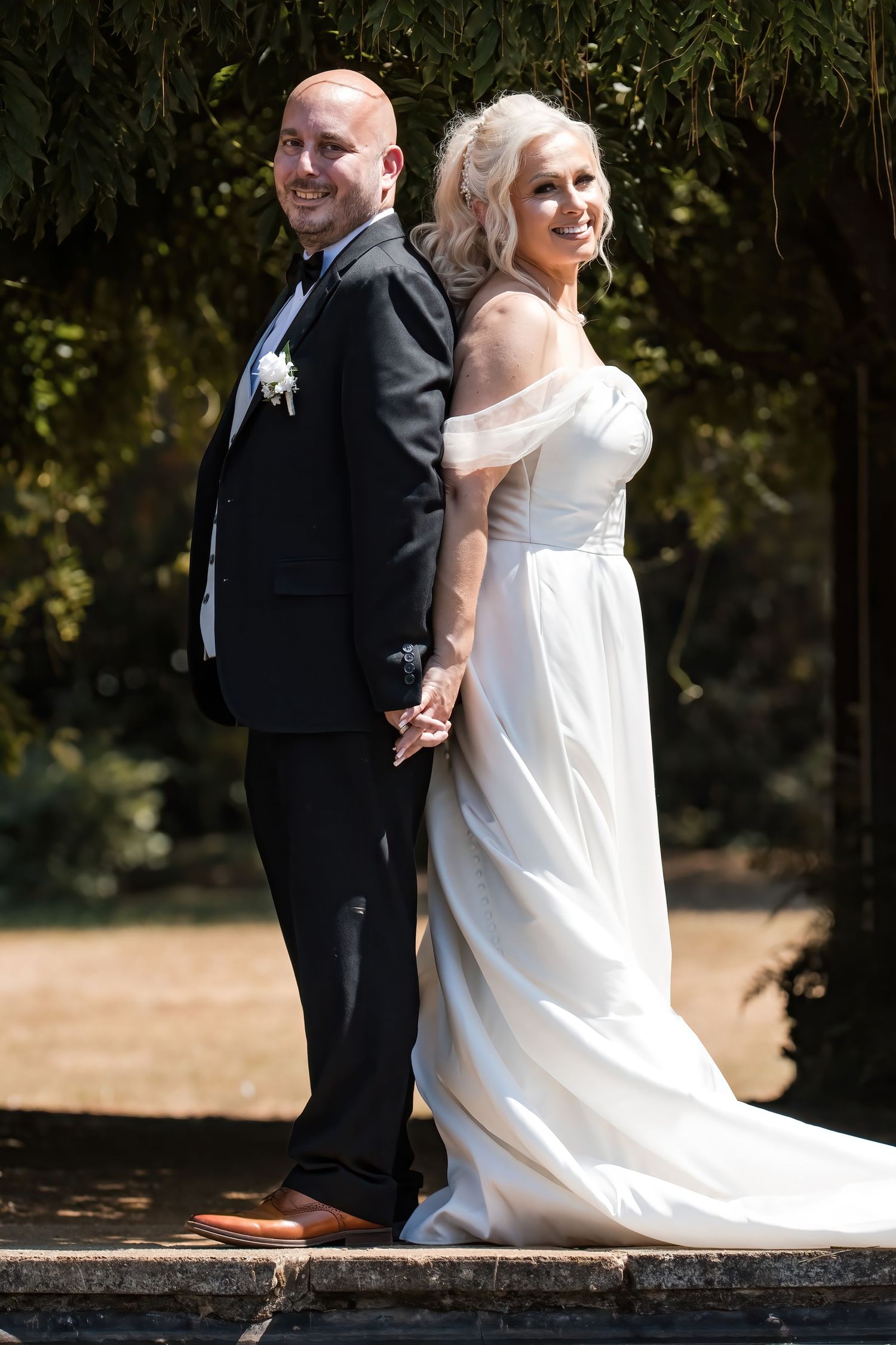 Smiling wedding couple standing back to back holding hands in sunlit garden, bride in flowing white dress.