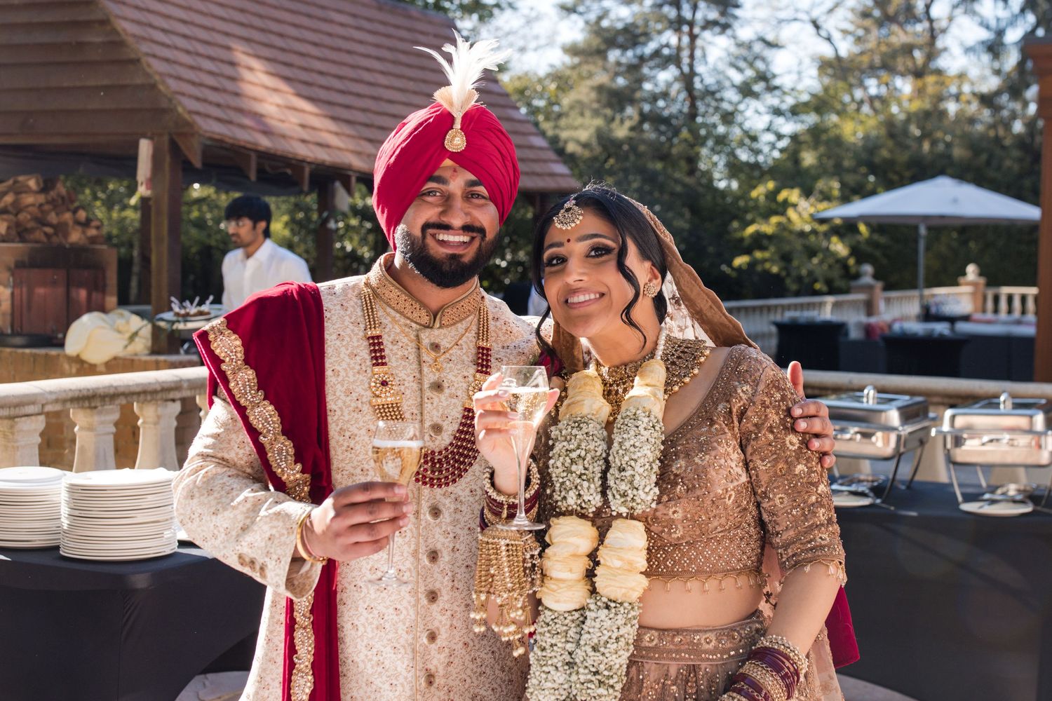 Sikh bride and groom in traditional gold and red wedding attire smile and raise glasses outdoors.
