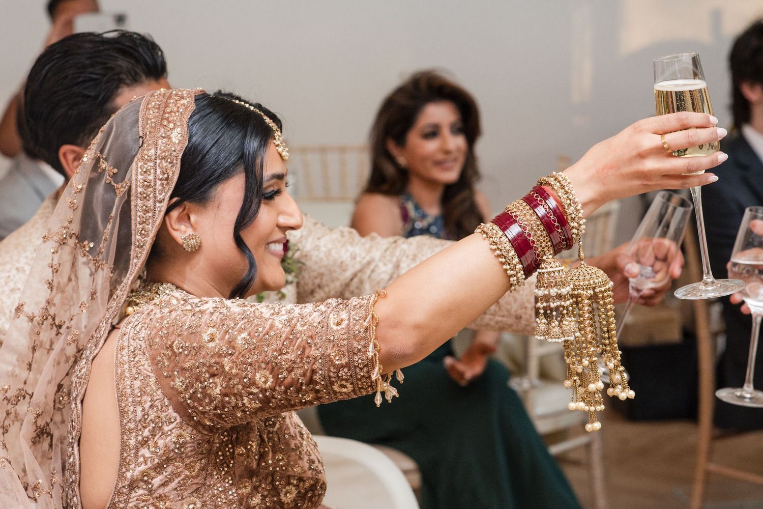 Indian bride in gold lehenga with red bangles raising champagne glass in a joyful wedding toast.