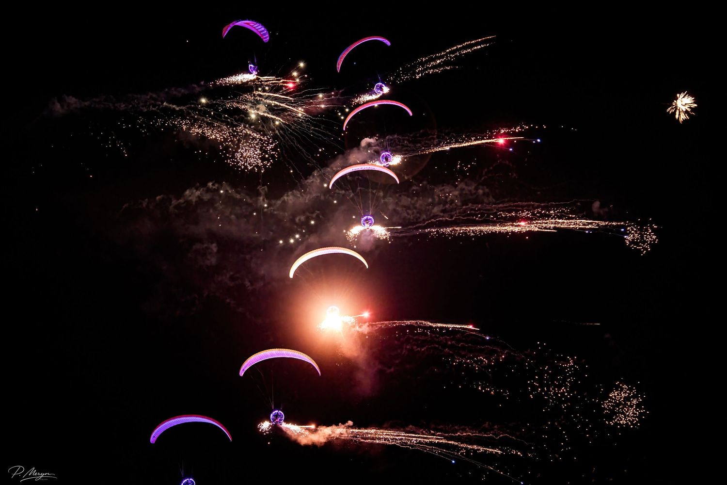 Illuminated paragliders soar through a dark night sky amid bursting fireworks and golden light trails.