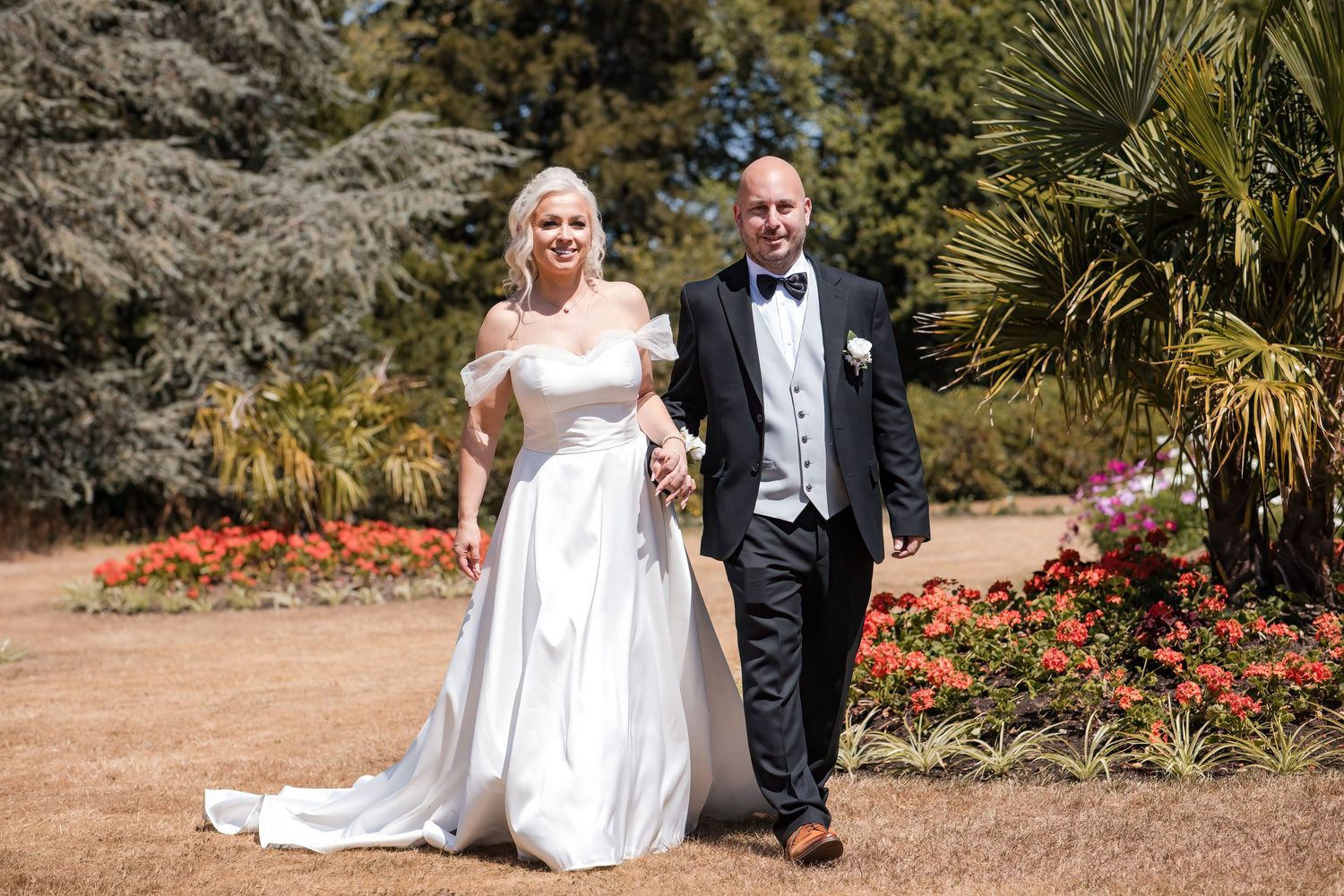 Smiling bride in white gown and groom in tuxedo walk hand in hand through a sunny garden.