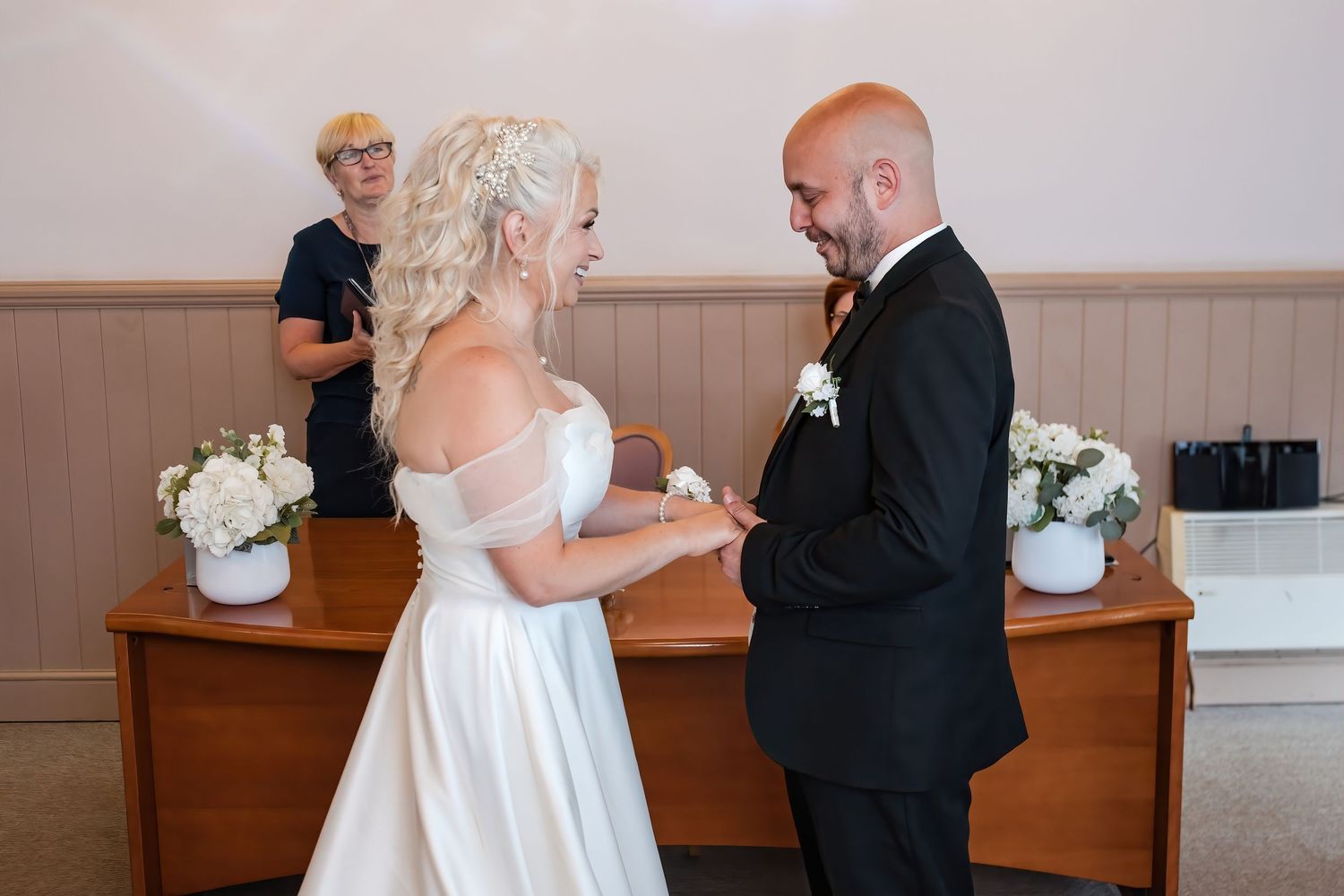 Bride and groom exchange vows at wedding ceremony with officiant standing behind wooden altar decorated with white flowers.