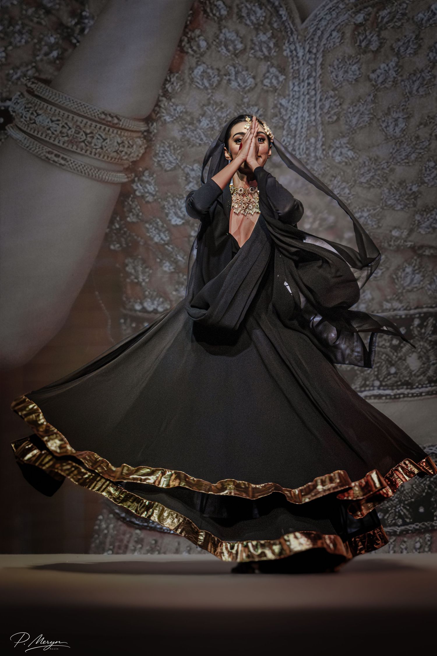 Model in black lehenga with gold border twirls dramatically during South Asian fashion show performance.