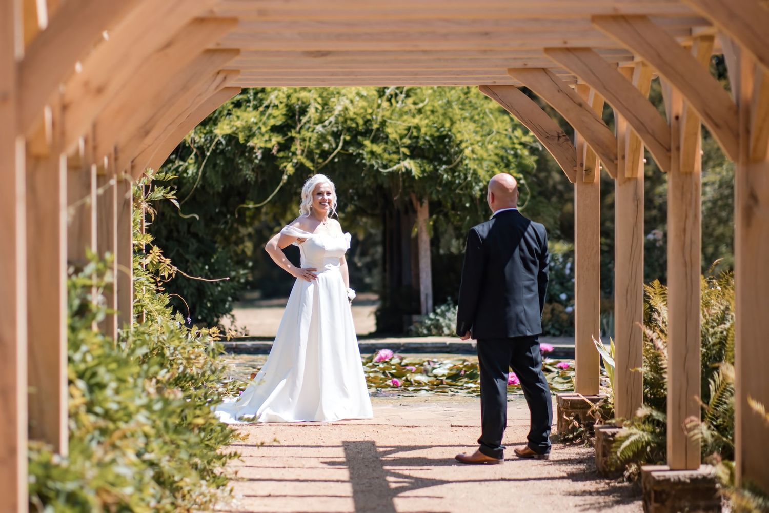 Bride and groom share a candid moment under a wooden pergola in a beautiful garden setting.