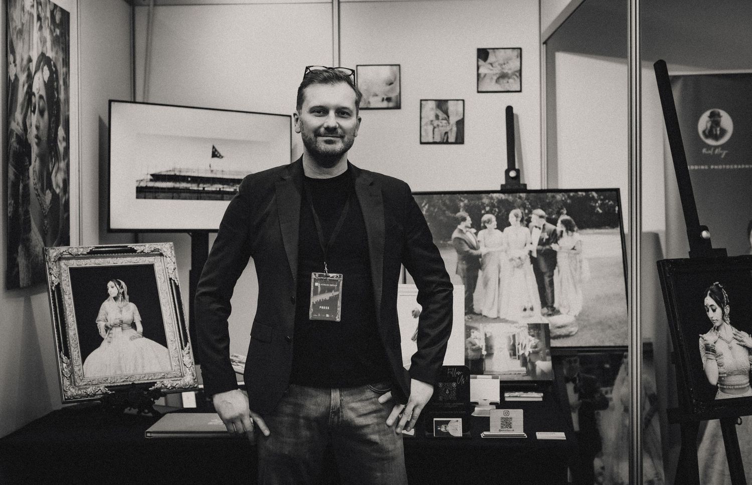 Man with beard standing confidently at an art exhibition booth displaying black and white photography prints.