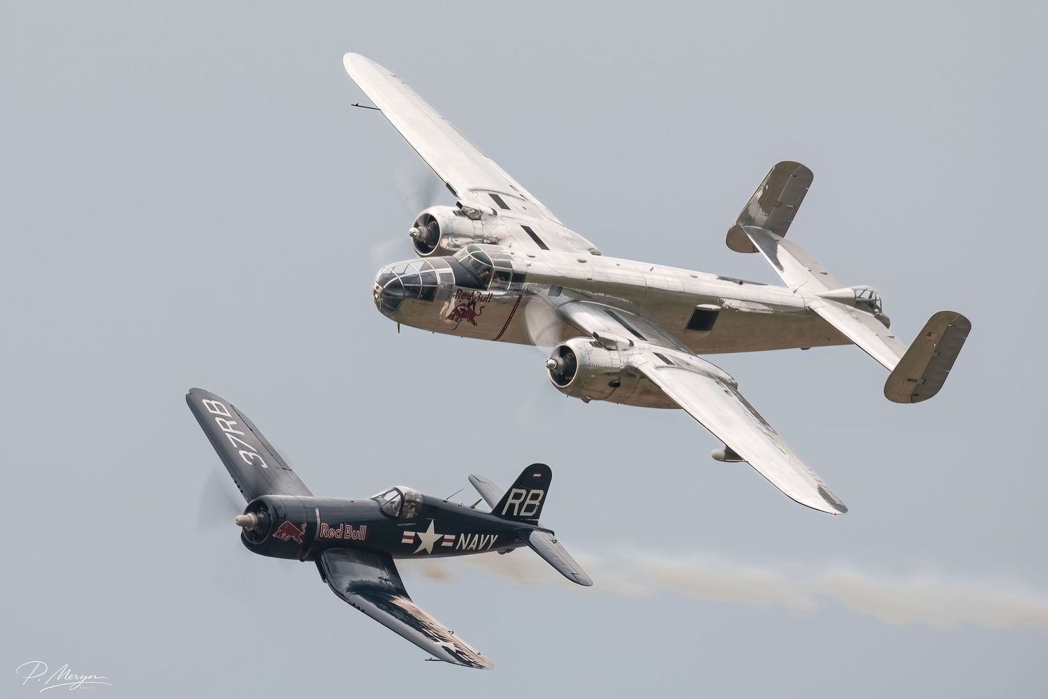 A vintage WWII B-25 bomber and Navy Corsair fighter fly in formation at an airshow.