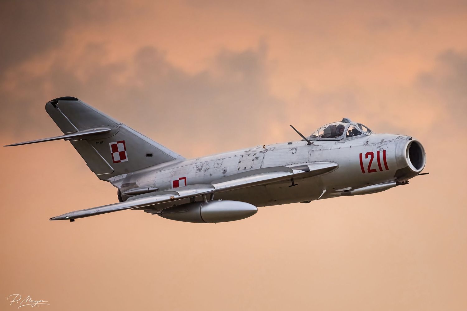 A Polish Air Force MiG-15 jet fighter flies against a dramatic orange sunset sky.
