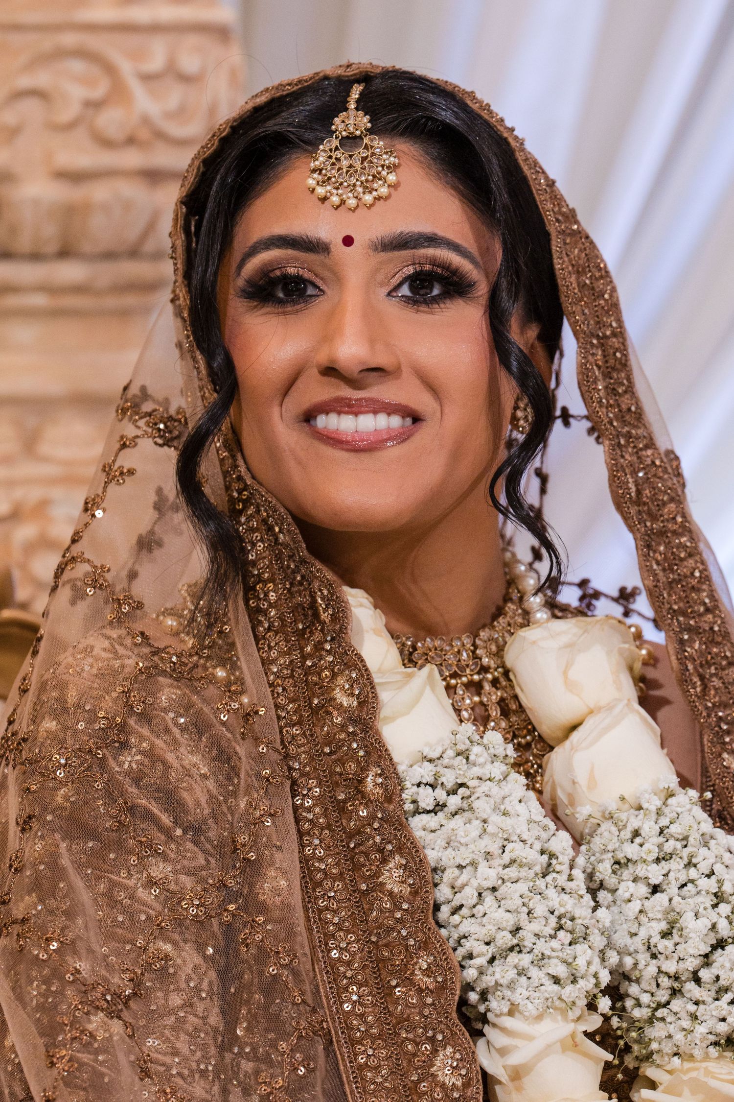 Beautiful Indian bride in gold embroidered lehenga smiles holding white rose bouquet at wedding ceremony.