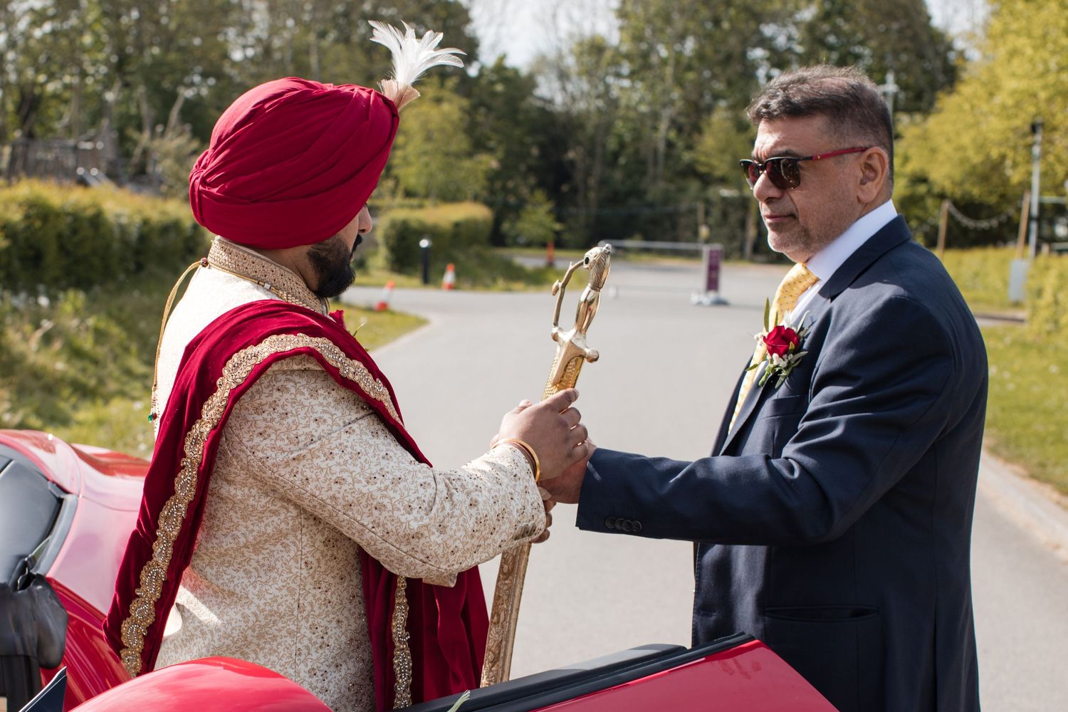 Sikh groom in ornate sherwani and red turban greets a suited guest beside a red car.