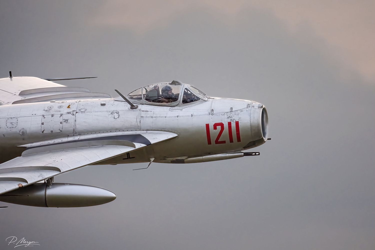 Close-up cockpit view of vintage MiG-15 aircraft numbered 1211 in silver finish flying through overcast skies.