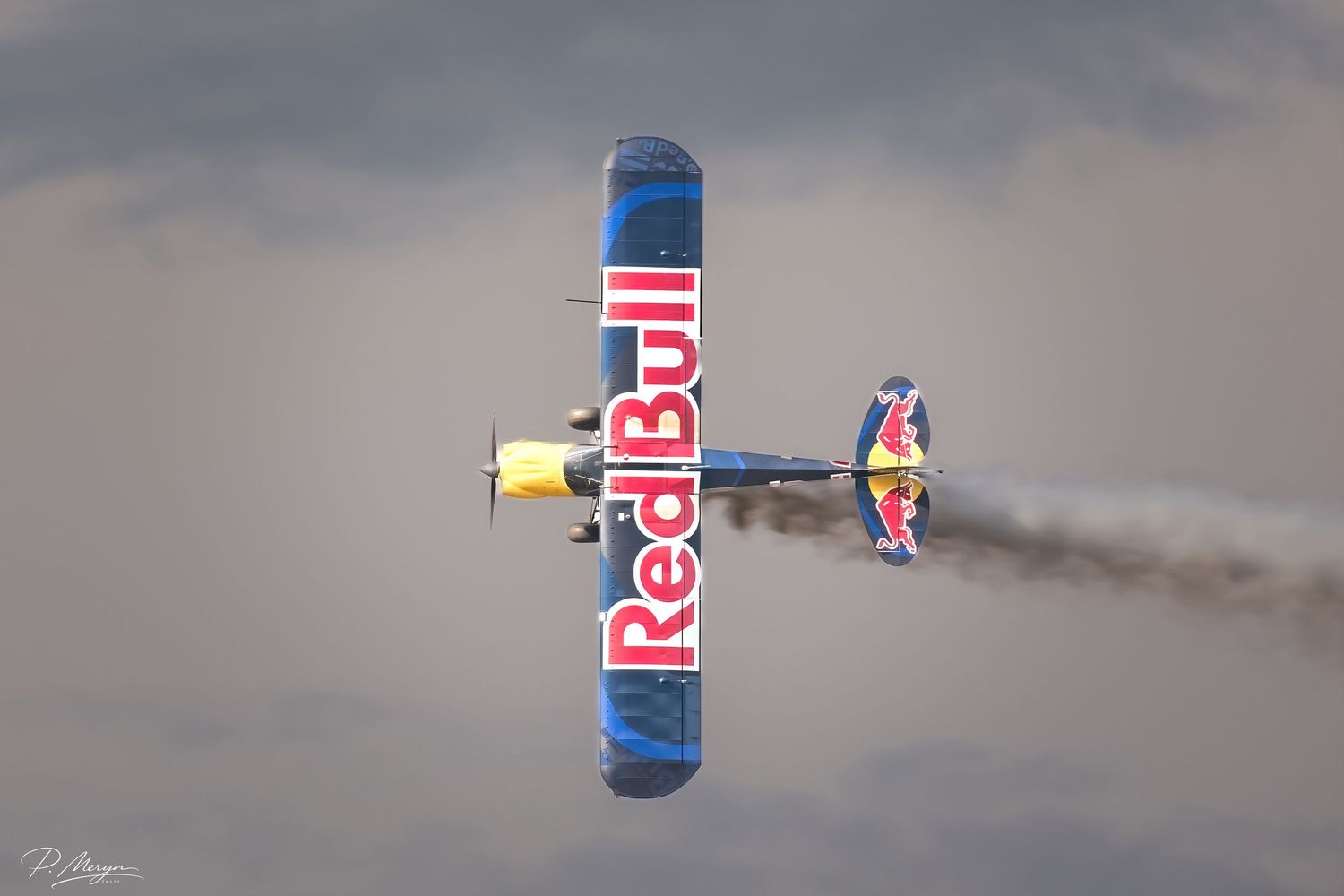 Red Bull branded aerobatic biplane performing inverted flight maneuver with smoke trail.