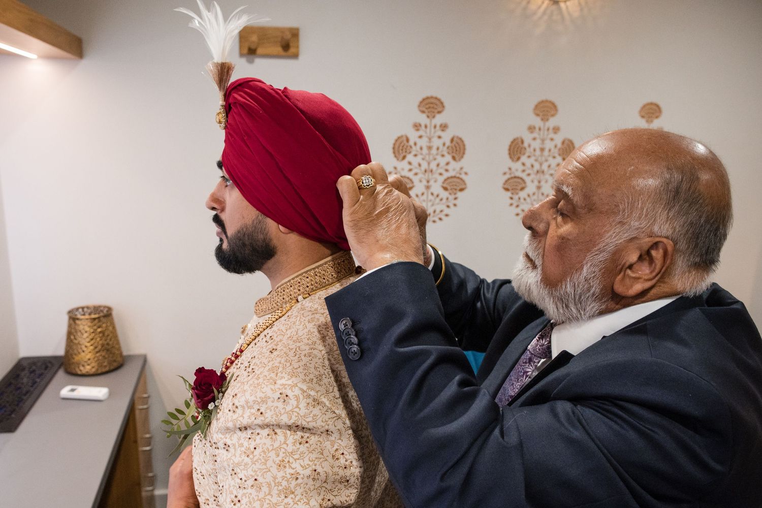 Elder man adjusts the red turban of a Sikh groom during traditional wedding preparations.