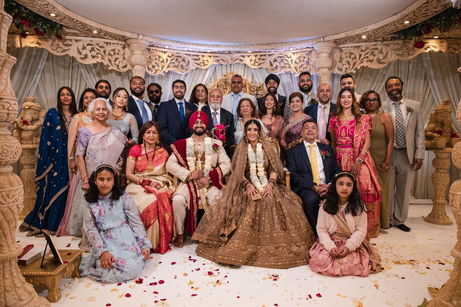 Large family group portrait with Sikh bride and groom seated at center in elaborate wedding venue.