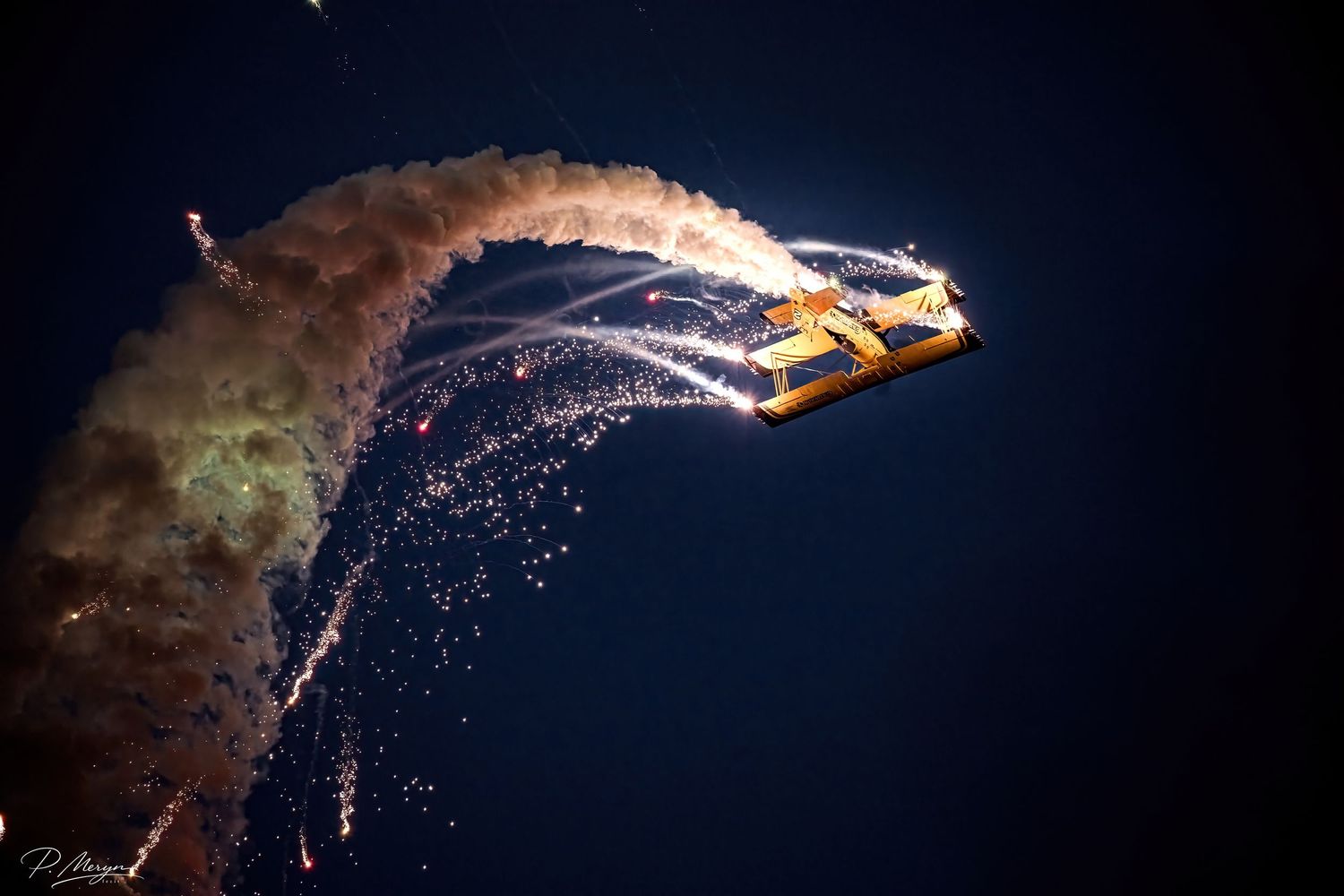 Yellow biplane performing stunning nighttime pyrotechnic display with sparks and smoke trails.