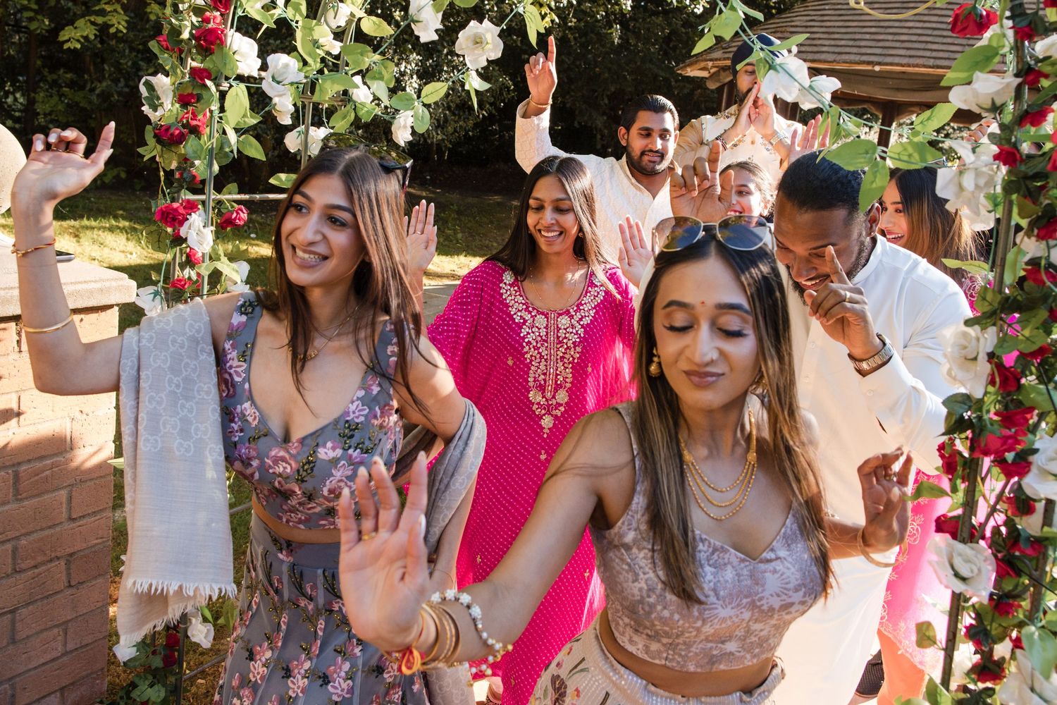 Smiling guests in traditional Indian and Western attire dance under a floral archway at a wedding.