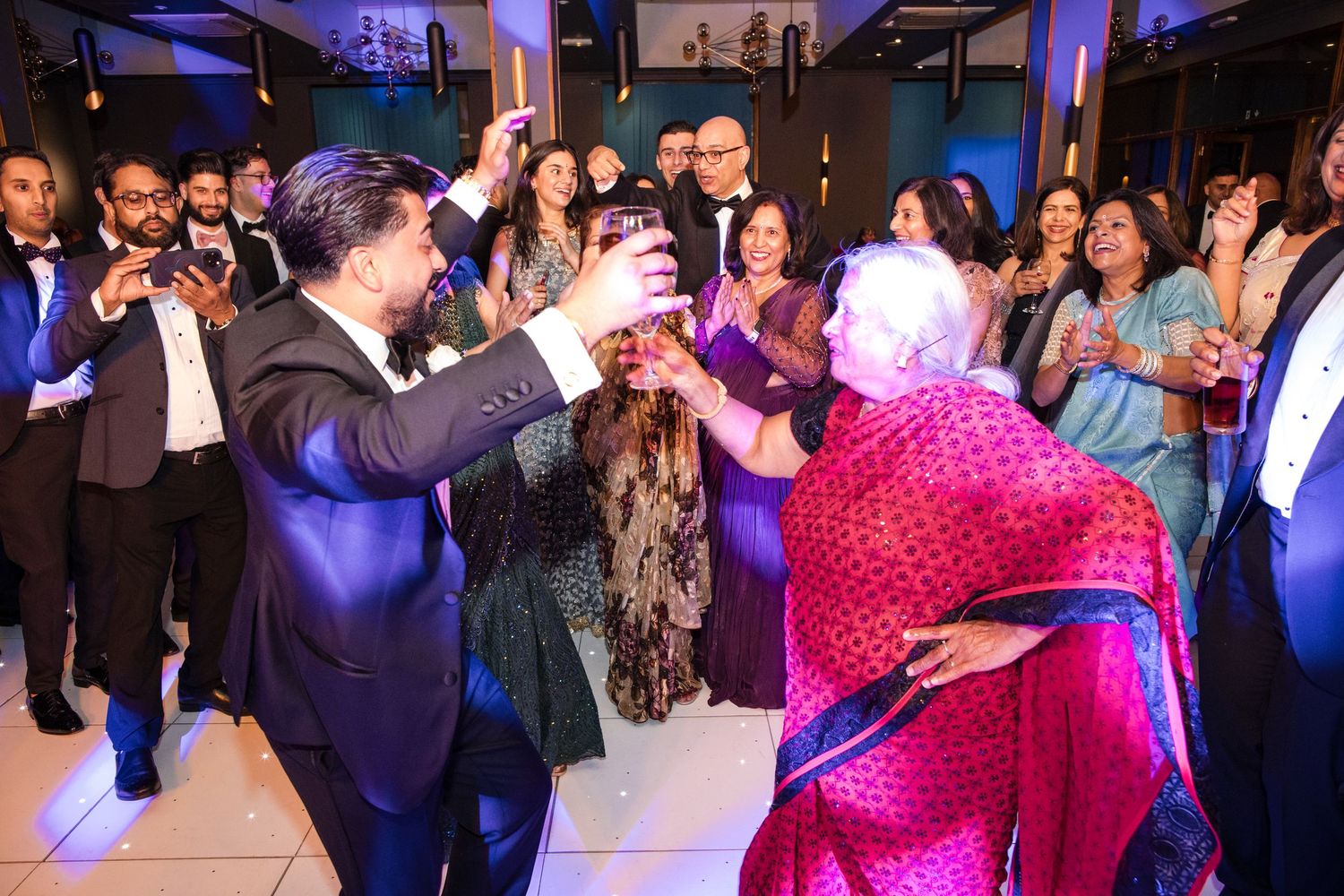Man in blue suit toasting drinks with elderly woman in pink sari at lively Indian wedding reception.