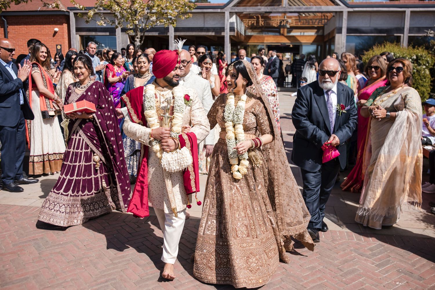 Sikh bride and groom in traditional gold and red attire walk together during outdoor wedding procession.
