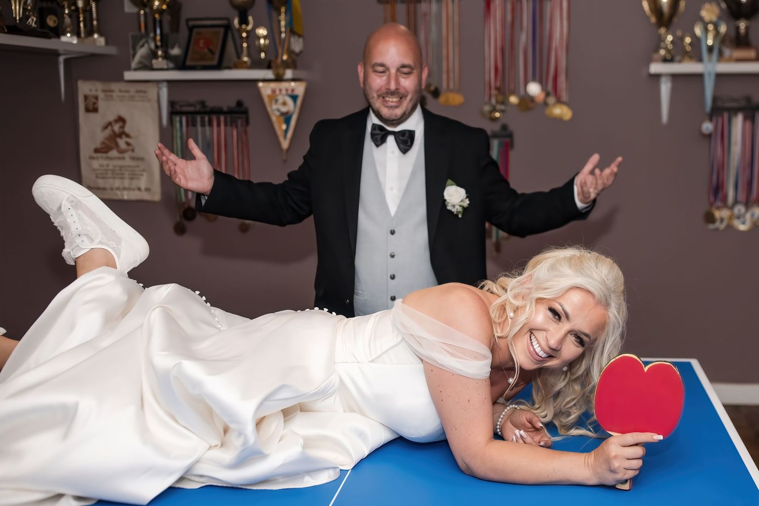 Bride poses playfully on ping pong table holding paddle while groom gestures behind her.