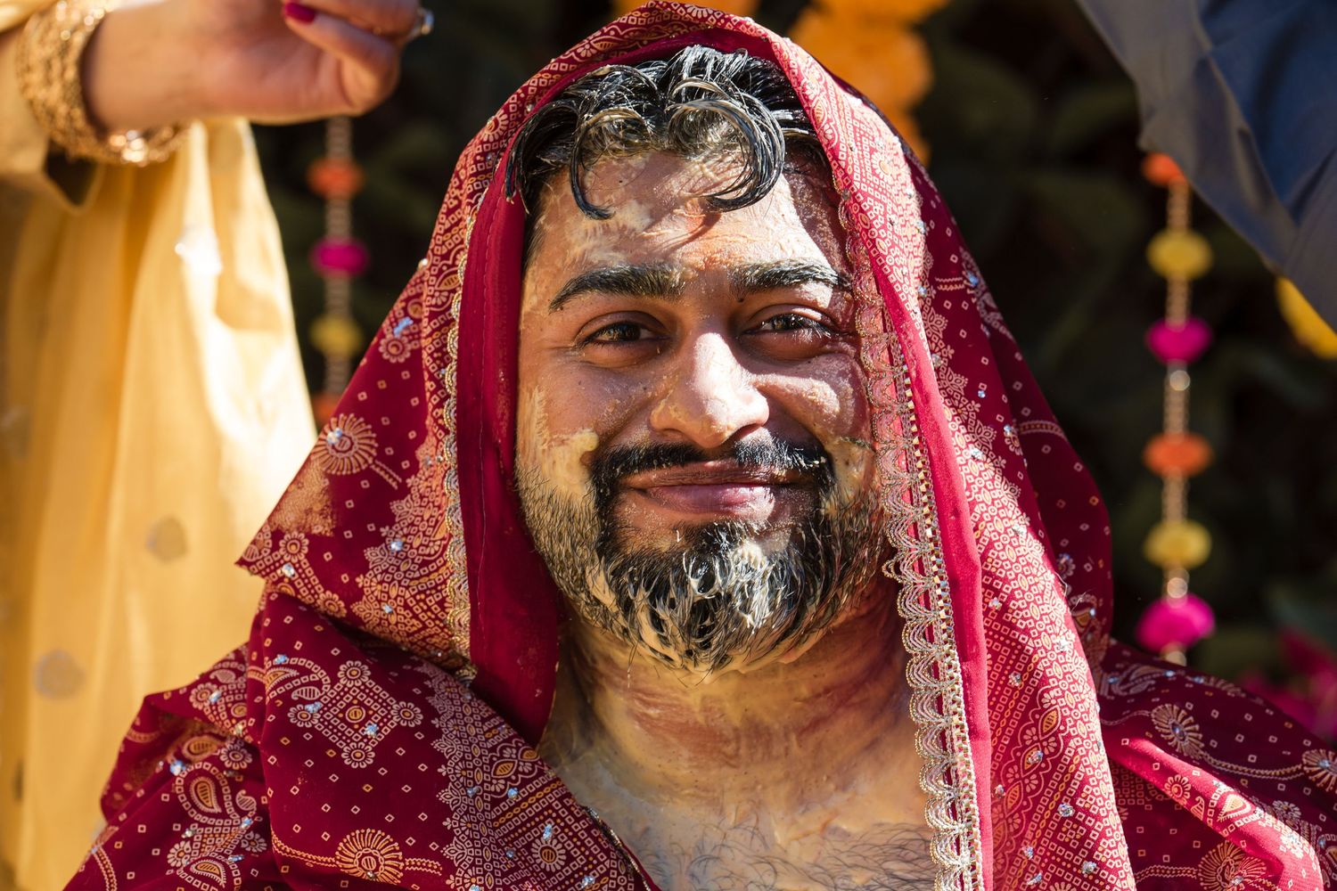 Smiling groom draped in red embroidered fabric with haldi turmeric on his beard at Indian wedding.